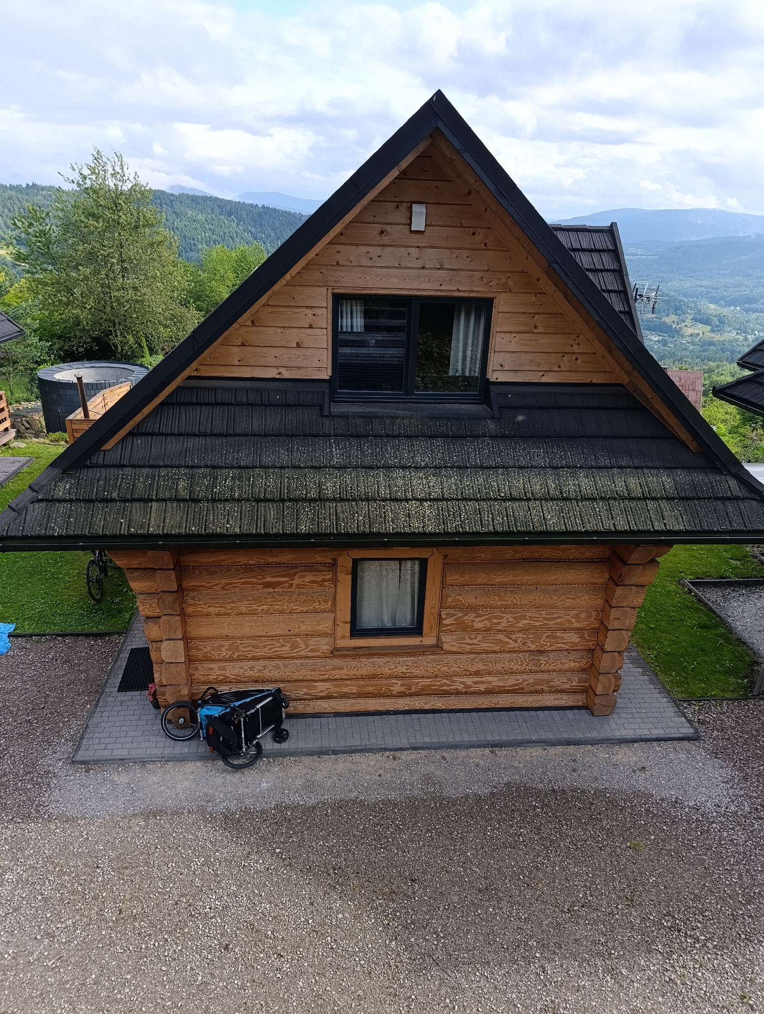The image shows a wooden cabin with a dark, tiled roof in Stróża, Poland. The cabin has a pitched roof and several windows. In front of the cabin, there is a paved walkway and a gravel area. A blue and black bicycle trailer is lying on its side on the walkway. To the left of the cabin, there is a round hot tub. The background shows a landscape of rolling hills covered in trees, with a cloudy sky above. The scene appears to be a tranquil, rural setting.