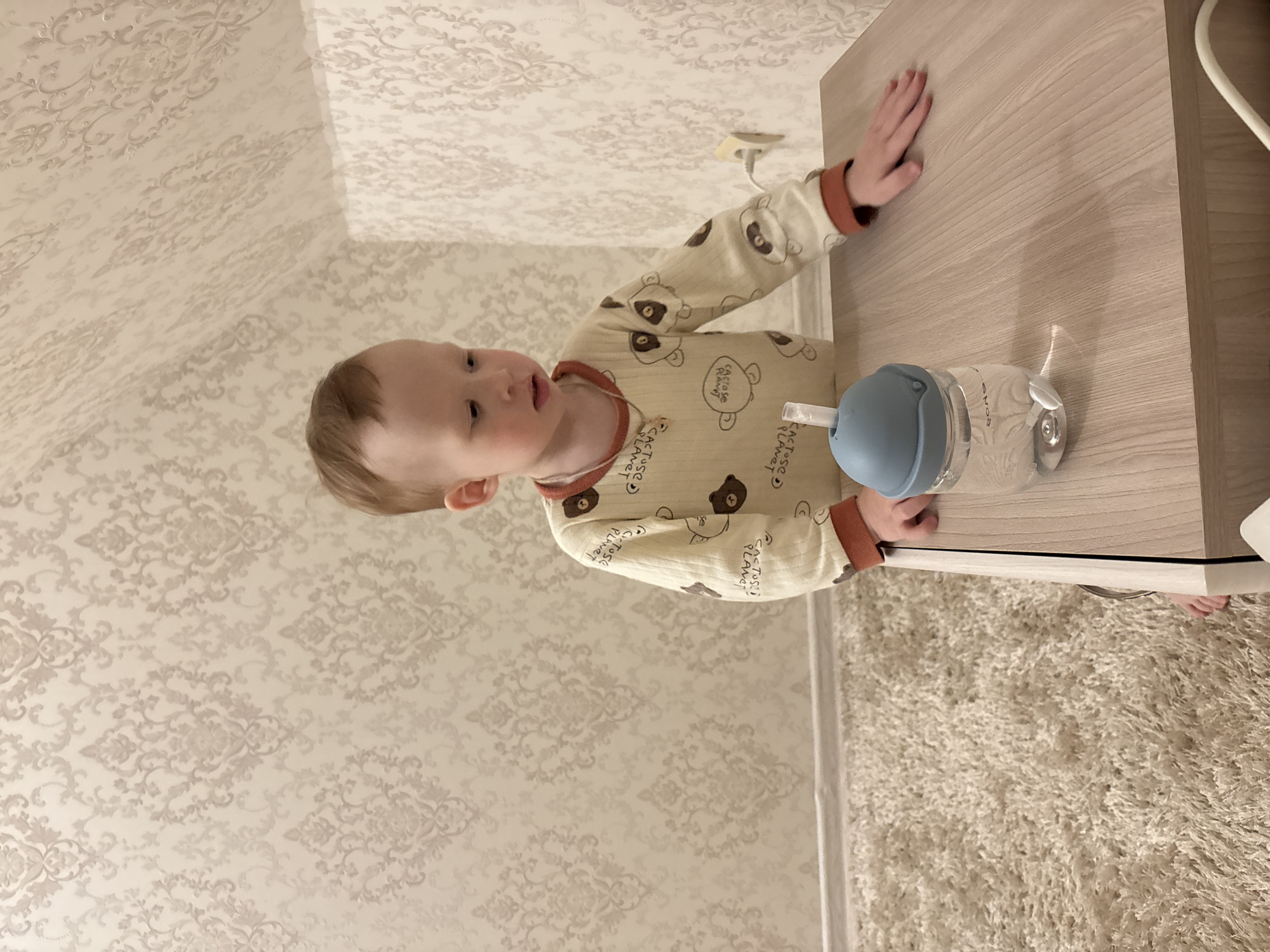 A toddler wearing a long-sleeved, light-colored pajama set with a pattern of cartoon bears is standing next to a light wood-grain surface, likely a table or countertop. The toddler is reaching for a blue and clear plastic water bottle with a straw. The background shows a patterned wallpaper and a section of carpet. The location is in a room of a building of undetermined location. The prompt specified Ferma, Russia, but it is impossible to verify this from the image.