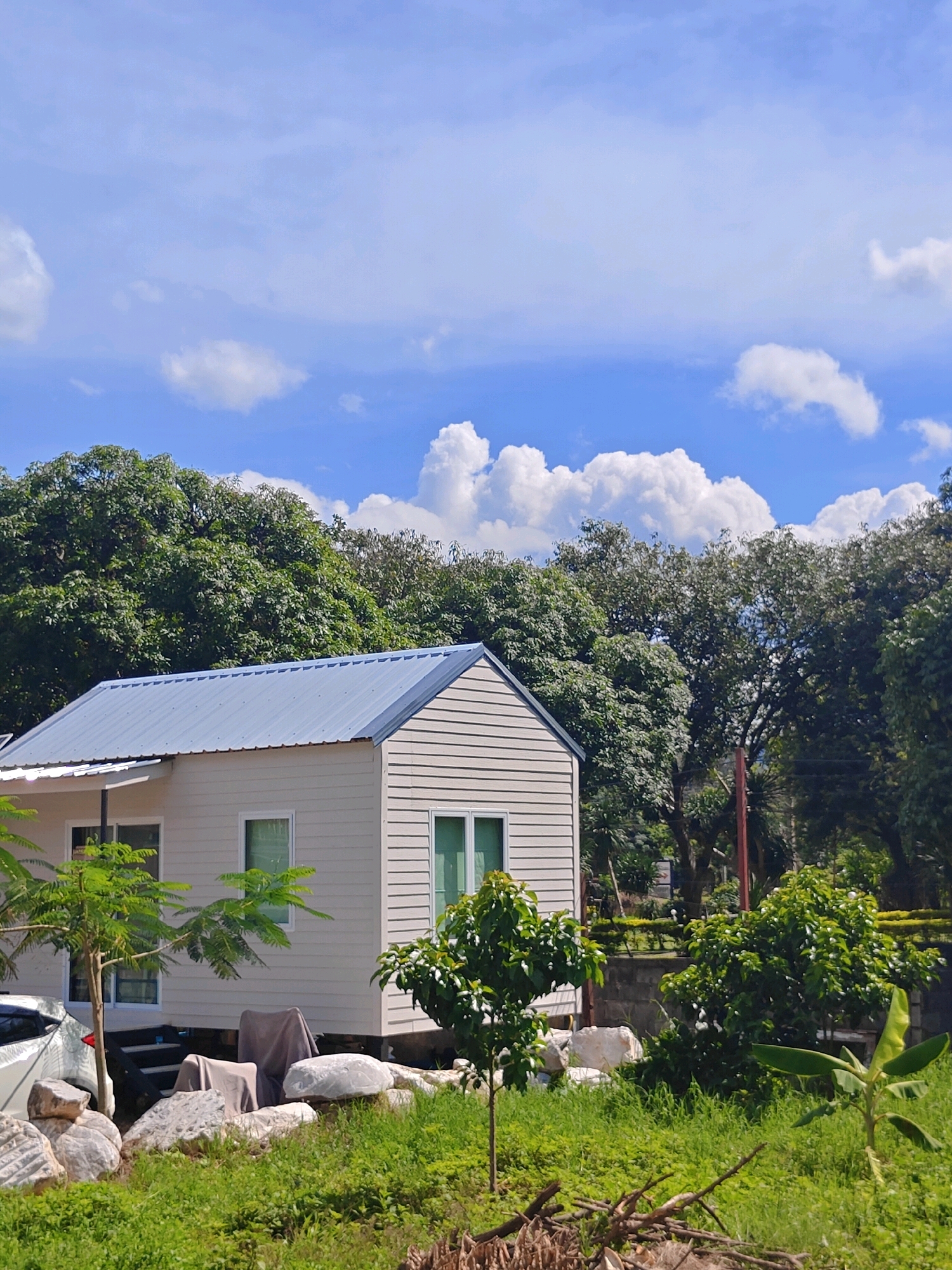 The image depicts a small, modern house with a grey metal roof, situated in a lush, green outdoor environment. The house features light-colored siding and several windows. In front of the house, there is a small tree, and beyond that, a white car is partially visible. The foreground consists of green grass, rocks, and some dried branches. The background is dominated by dense, green trees, and a bright blue sky with scattered white clouds. The scene suggests a rural or semi-rural setting, possibly a residential area outside of a major city, consistent with the location of Khao Yai, Thailand, known for its natural beauty and resorts. There are no people visible in the image, and the overall atmosphere is serene and natural.