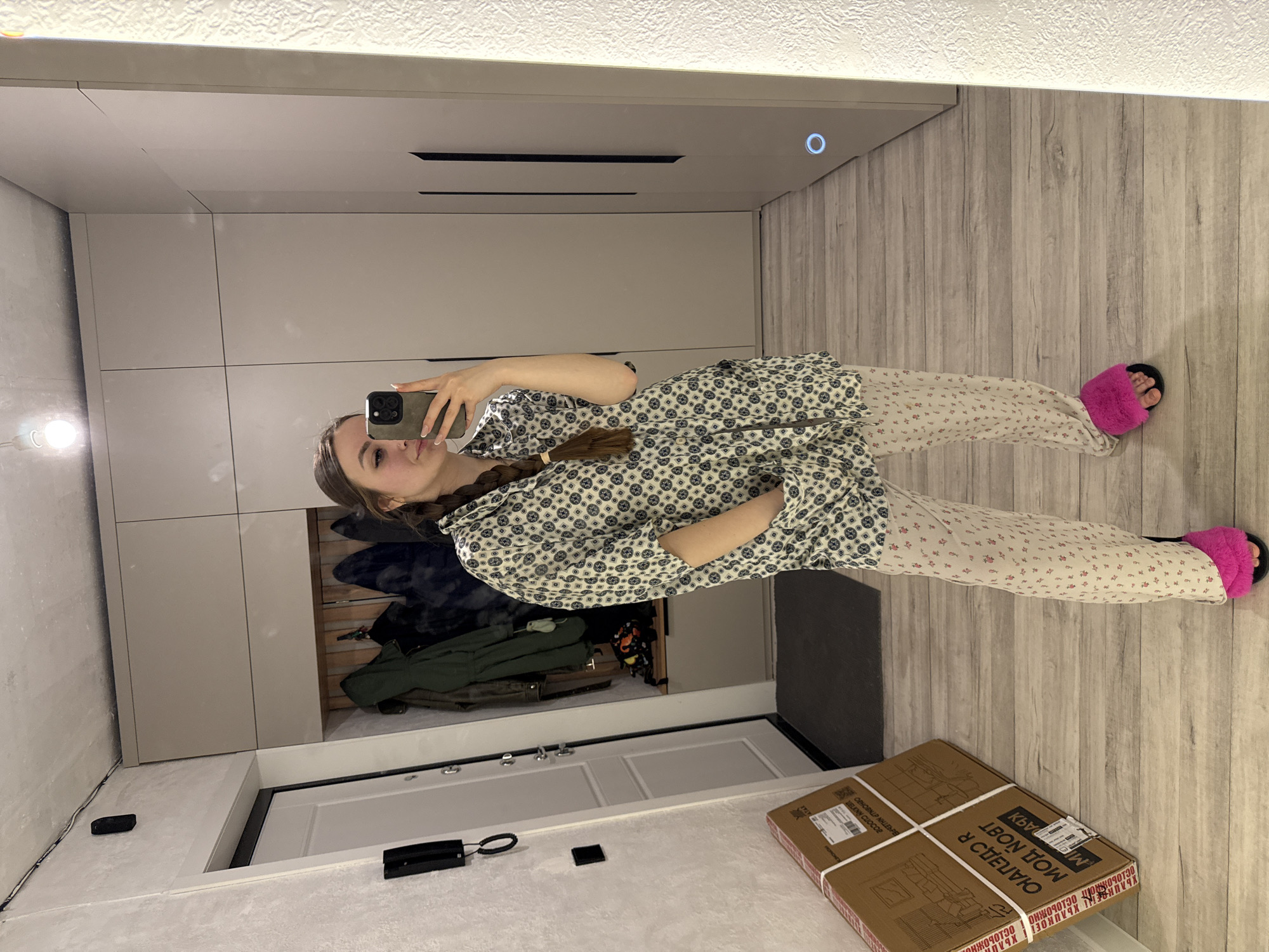 A woman stands in front of a mirror, taking a selfie. She is wearing a patterned pajama set and pink slippers. The mirror reflects a closet with clothes hanging inside, a white door, and a cardboard box on the floor. The room has light-colored walls and wood-like flooring. The location is Unknown, Unknown.