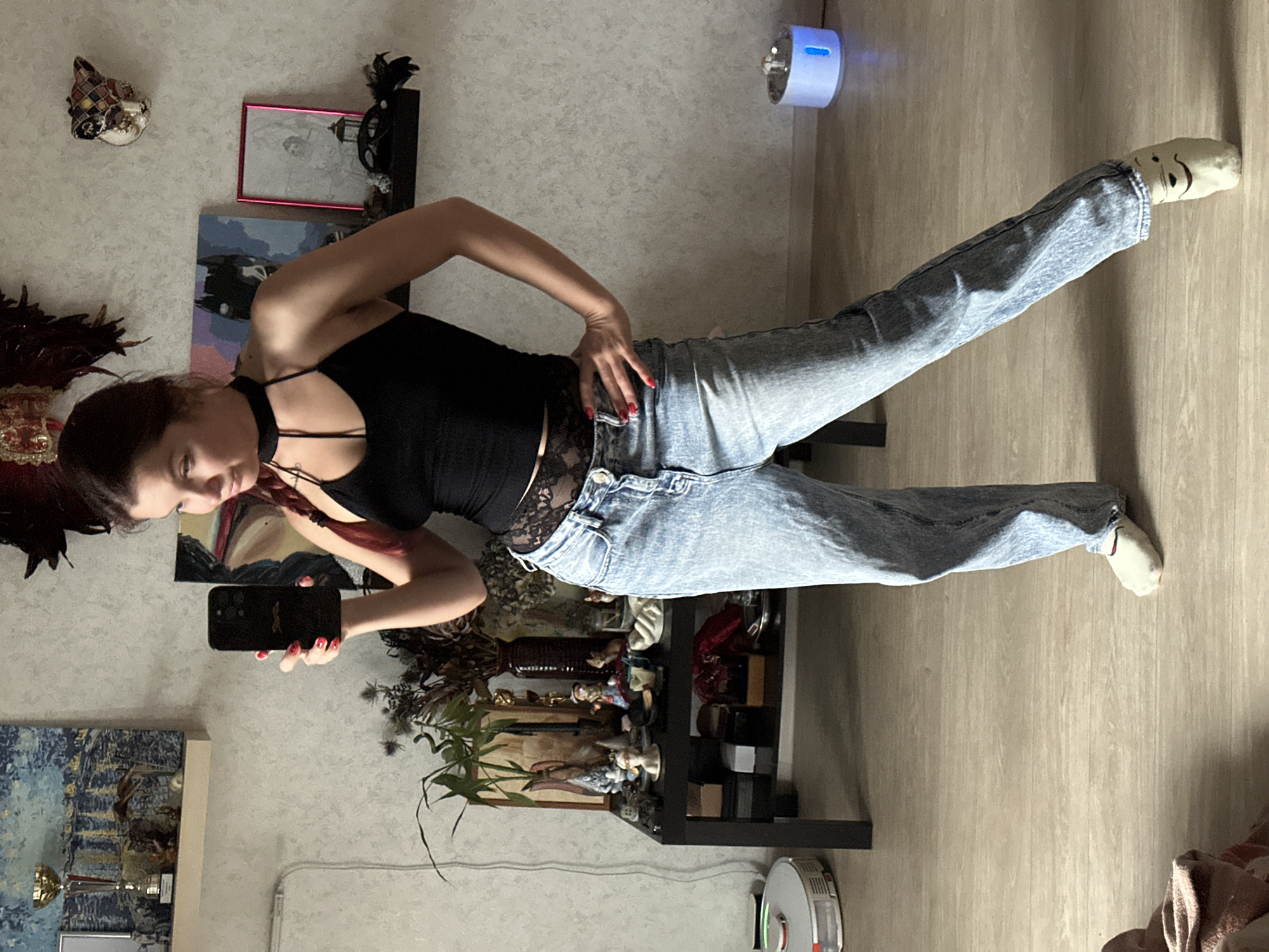 The image depicts a young woman taking a mirror selfie in an indoor setting, possibly a residential apartment. She is wearing a black tank top with lace detailing at the waist, high-waisted light-wash denim jeans, and white socks with cartoon faces. She has her hair in a braid. Behind her, a black table is cluttered with various decorative items, including vases, figurines, and plants. Shelves on the wall above the table display more objects, such as a trophy and artwork. Other elements in the room include a robot vacuum cleaner on the floor to the left and a blue illuminated object to the right. The overall scene suggests a casual, personal space. There is no discernible information about the city of Vladimir, Russia, or any other specific location.