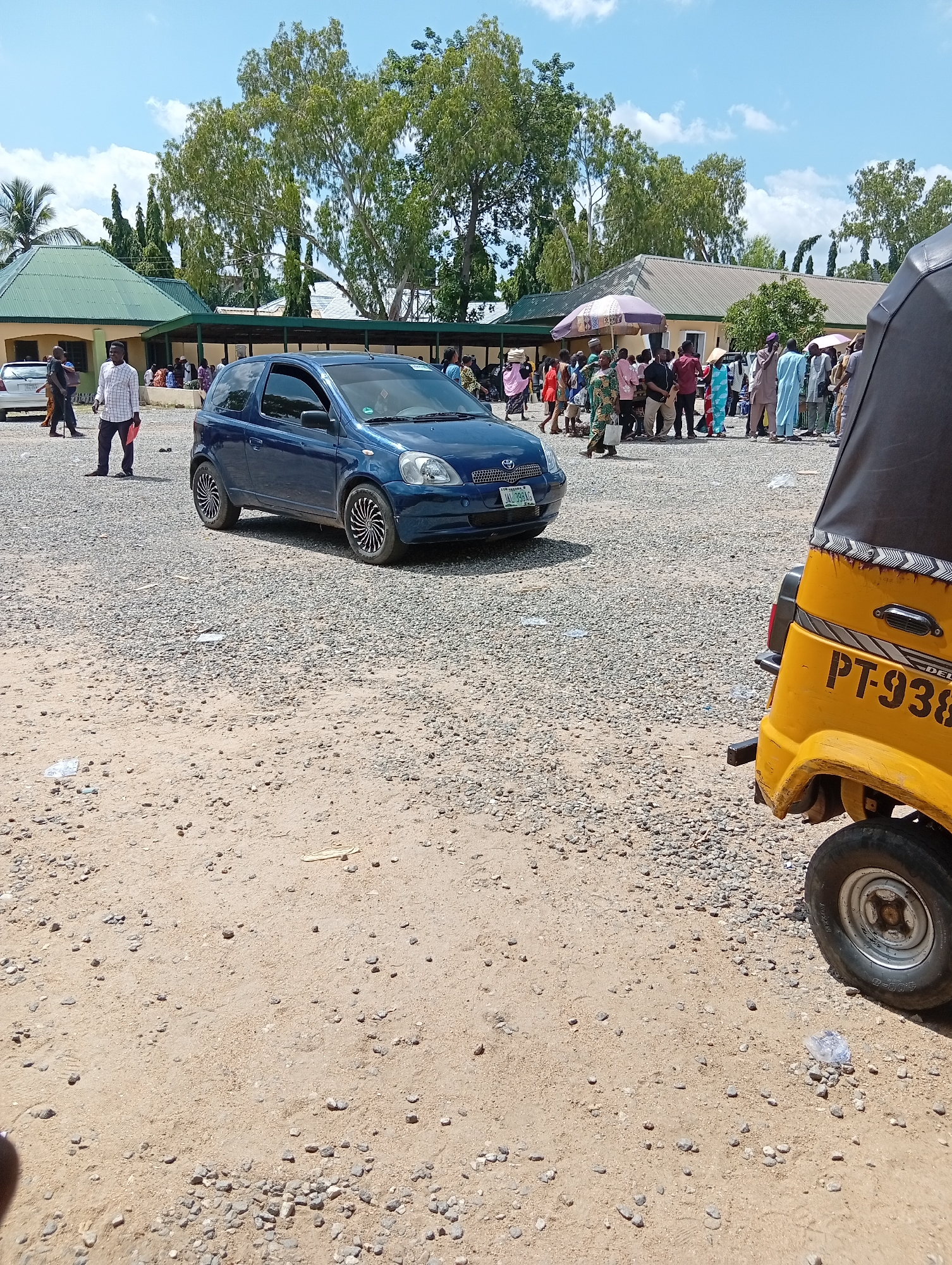 A blue hatchback car is parked on a gravelly dirt lot in front of a low-rise building with a green roof. Several people are gathered near the building, some standing under umbrellas. To the right of the blue car, the rear of a yellow three-wheeled vehicle is visible, with the license plate "PT-938" printed on its side. The scene appears to be outdoors, with trees and a blue sky with clouds visible in the background. The overall impression is of a public gathering or event taking place in a dusty, open area in Jalingo, Nigeria.