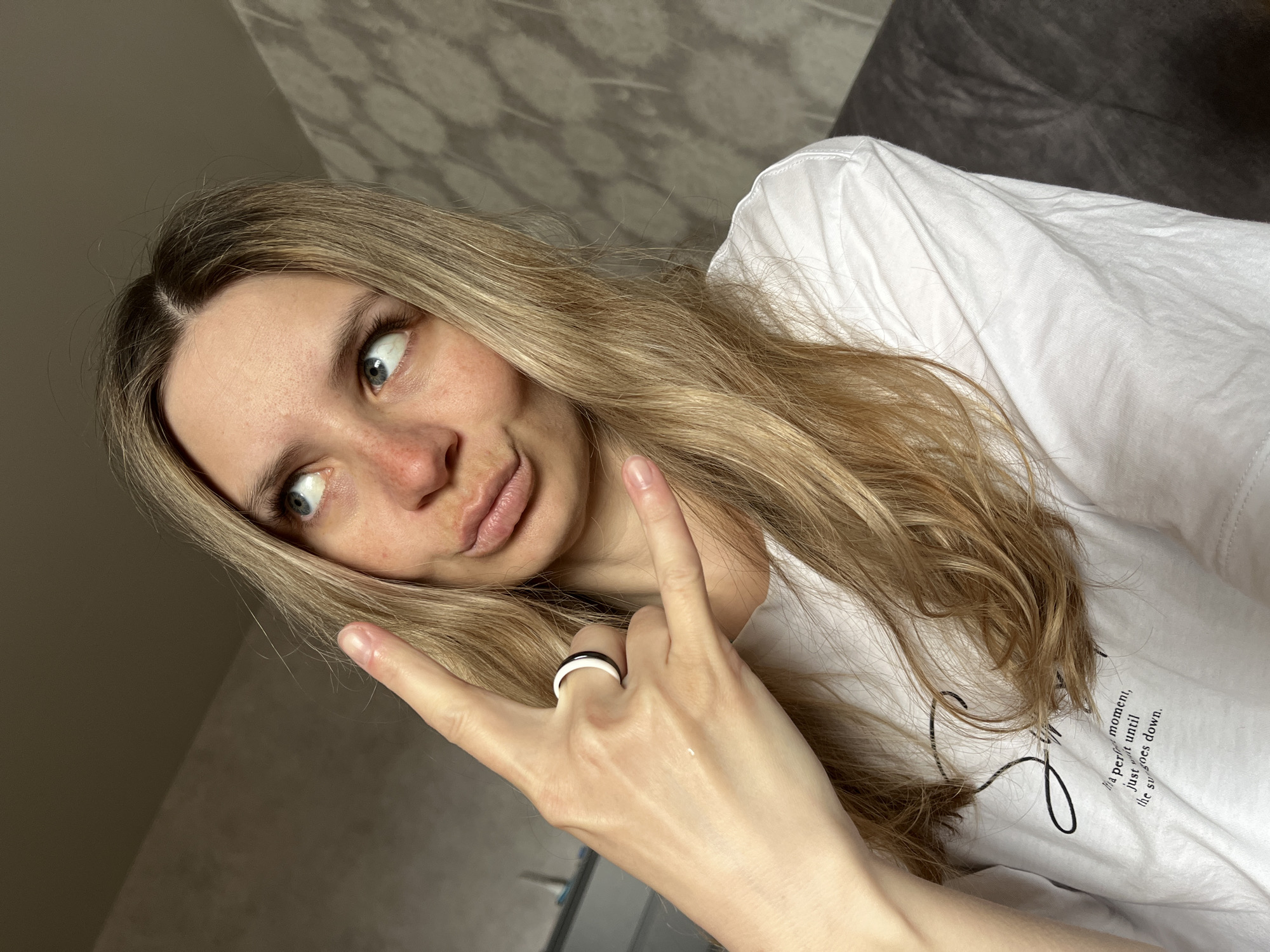 A blonde woman with light skin and green eyes is posing for a selfie. She's wearing a white t-shirt and making a "rock on" hand gesture. A ring with a white and black band is visible on her finger. The background includes a patterned wall, a gray surface, and a piece of furniture. There is no indication that this image was taken in Chaikovsky, Russia.