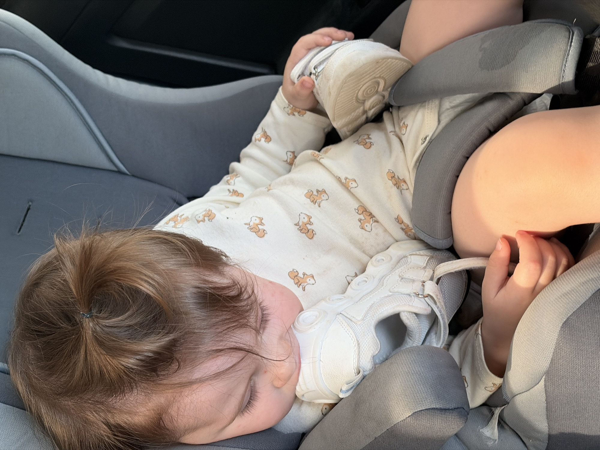 A young child, with brown hair pulled into a small ponytail, is sleeping soundly in a grey car seat. The child is wearing a white pajama set adorned with a pattern of orange foxes. One of the child's feet is resting on a white sneaker. The car seat appears to be in a vehicle, with dark interior visible in the background. The overall scene is one of peaceful slumber within a typical car setting. The location in the city of Ваньки, Russia is not determinable from the image.