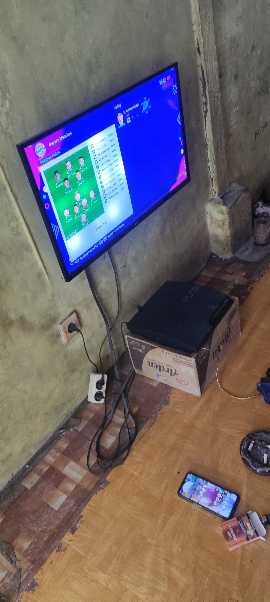 The image depicts an indoor scene, likely a living space, in Karangwangun, Indonesia. A flat-screen television is mounted on a wall, displaying a soccer game interface with player information and team selection. Below the television, a PlayStation console is placed on top of a cardboard box. Electrical cords connect the television and console to wall outlets. The floor is made of wooden parquet tiles, showing some wear and tear. A mobile phone and a pack of cigarettes are on the floor near the console. The walls appear to be textured and painted. The lighting is dim, suggesting it may be evening or a poorly lit room.