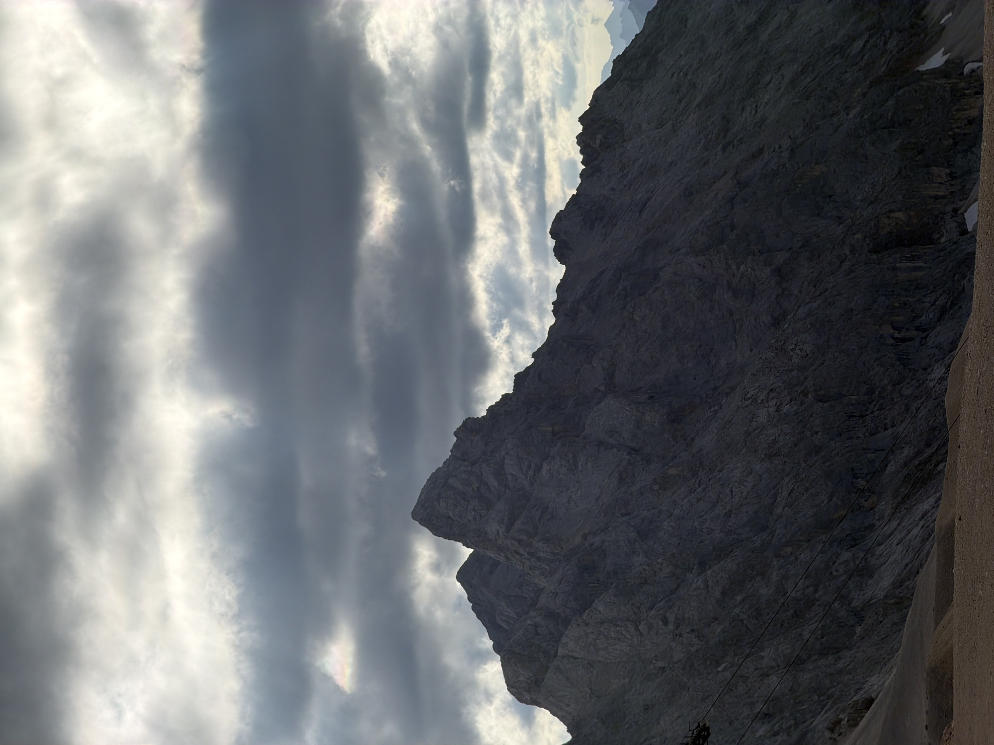 The image shows a rugged mountain range under a dramatic, cloudy sky. The foreground features a rocky slope leading up to the dark, textured mountainside, which is cast in shadow. Two prominent peaks are visible, with the sky above filled with layers of gray and white clouds, some illuminated by an unseen light source, creating a sense of depth and atmosphere. The lower left portion of the image shows a cable car structure and its cables, suggesting a tourist or transport facility in the mountainous terrain. The overall scene conveys a sense of imposing natural beauty and possibly challenging weather conditions. This is likely a view of the Zugspitze, Germany's highest peak, located near Garmisch-Partenkirchen.