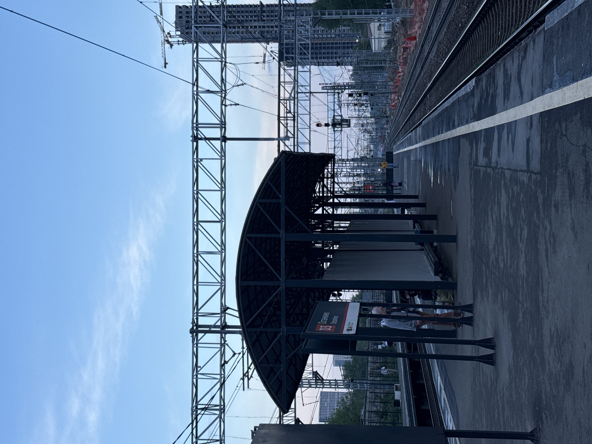 The image shows a train platform on the D3 line, specifically at the Ostankino station. Overhead power lines and support structures are prominent above the platform. A partially covered waiting area with signage is visible. In the background, railway tracks extend into the distance. One or two people are on the platform. A construction crane and a partially constructed building are present in the distance. The sky is light blue with some wispy clouds.