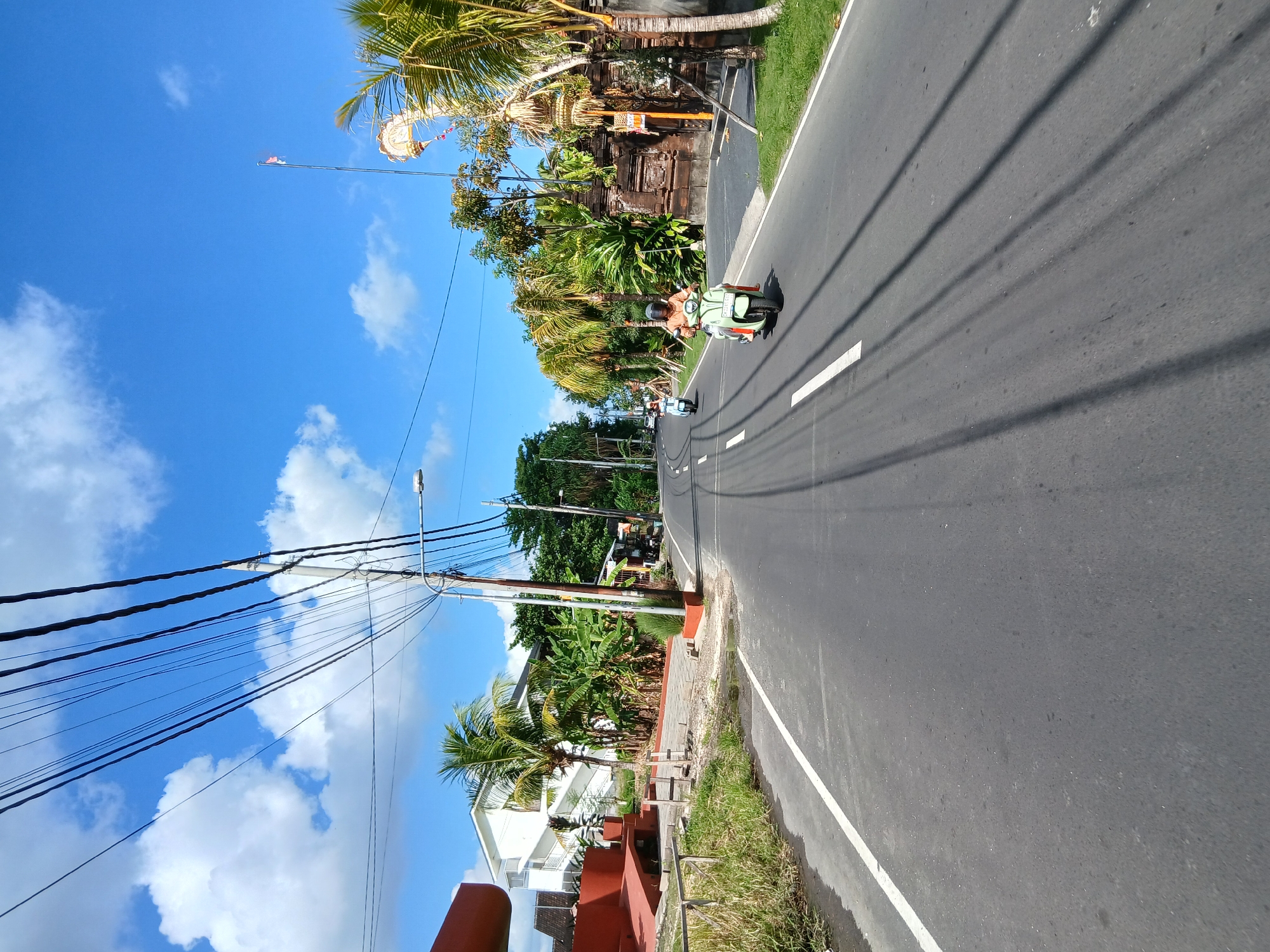 The image captures a sunny day on a paved road in Mengwi, Indonesia, with a clear blue sky dotted with fluffy white clouds. The road is marked with white lines and leads into the distance. On the right side of the road, there is lush green vegetation, including palm trees and dense foliage, behind a traditional Balinese stone wall. A flag flutters on a tall pole. Further down the road, a person is riding a light green scooter, wearing an orange shirt and a helmet. Another scooter, lighter in color, is visible further ahead. On the left side of the road, there is a modern white building with stairs, accompanied by tropical plants and a red-colored structure. Overhead, a dense network of black electrical wires crisscrosses the sky.
