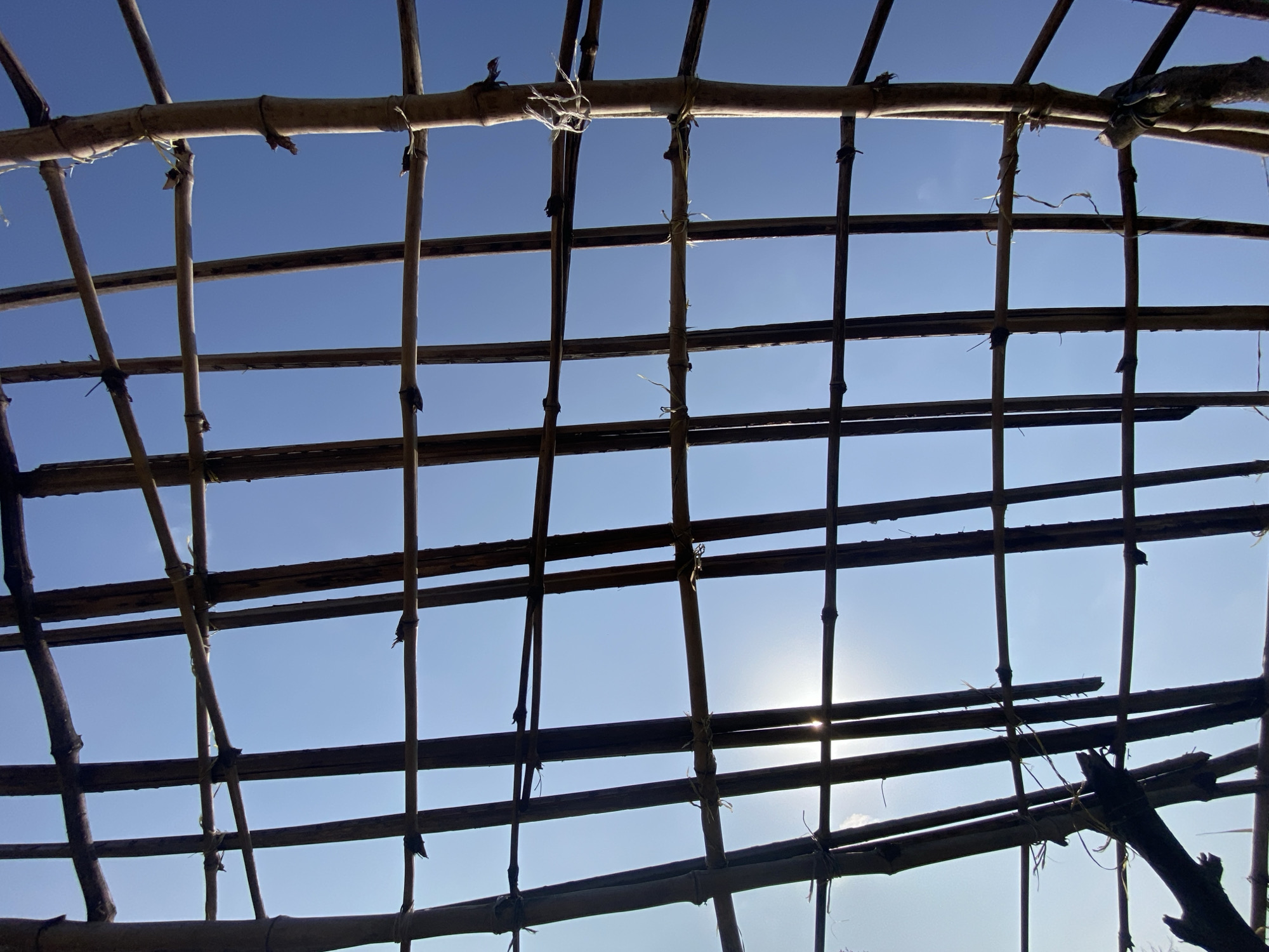 This image captures a close-up view of a bamboo-constructed roof framework against a clear blue sky. The bamboo poles, varying in thickness, are tied together with rough twine, creating a grid-like pattern. Some poles are positioned horizontally, forming the main support beams, while others are vertical or angled, intersecting to create the structure. The sunlight filters through the gaps, casting subtle shadows and highlighting the texture of the bamboo. There are no people or animals visible in the scene. The environment appears to be outdoors, with the bright sky suggesting daytime. The construction method and materials indicate a rustic or temporary shelter, potentially in a rural or less developed area. No specific landmarks or features are visible to pinpoint a precise location within the city of Ufu Esioni, Nigeria.