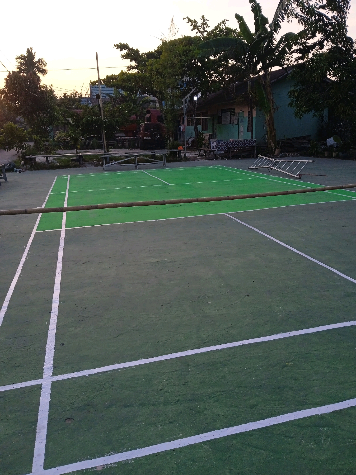 An outdoor tennis court in Banjarmasin, Indonesia, is pictured at dusk. The court is a hard surface, with green painted sections and white lines marking boundaries. A bamboo pole stretches across the court, suggesting it might be used for badminton or volleyball. In the background, trees and a light-blue building are visible, along with a ladder and some parked vehicles. The sky is clear and shows signs of sunset.