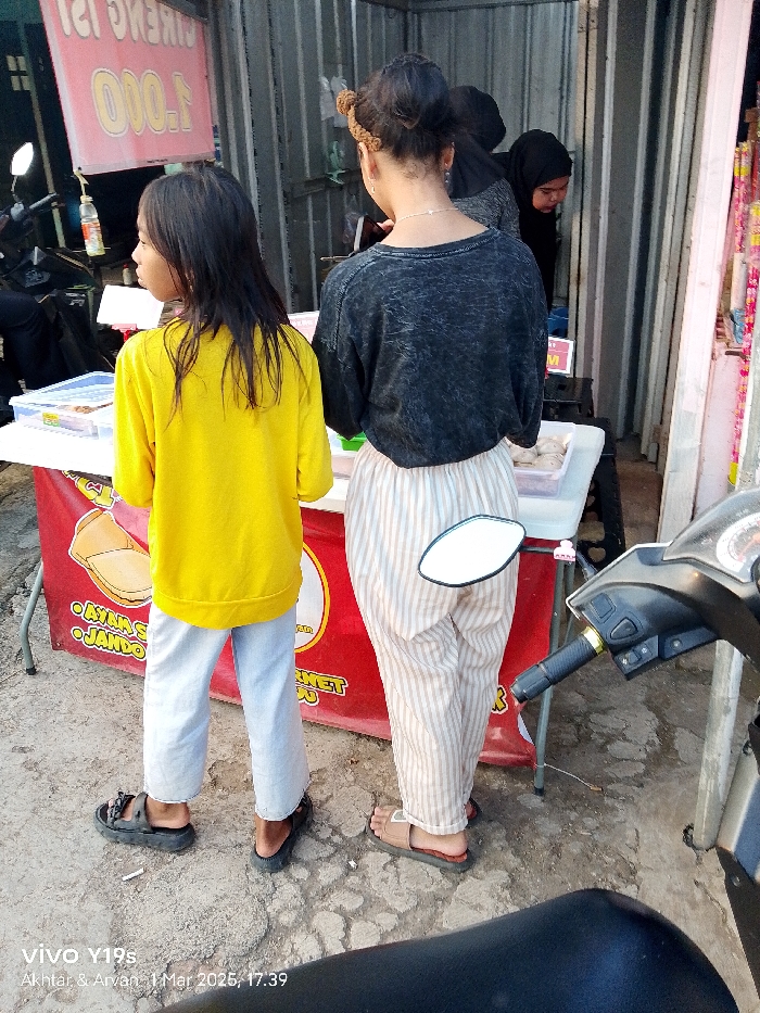 Two young females stand in front of a food stall, located in Kecamatan Haurgeulis, Indonesia, at coordinates -6.421 latitude, 107.996 longitude. The girl on the left is wearing a yellow shirt, blue jeans, and black sandals. She has long, dark hair and is looking to the left. The girl on the right is wearing a grey shirt, striped white pants, and black sandals. She has long, dark hair with a bun and a headband. She is looking down at the ground. A third female wearing a black hijab stands behind the girl on the right, looking at a stall displaying a food item. A black motorcycle is partially visible on the right side of the image, with a reflective rearview mirror. The food stall is decorated with red and white signs. A white table with a black food container sits in front of the stall. The ground is a mix of grey and brown concrete. A sign with the price "1.000" is visible on the wall to the left. The photo is taken from a low angle. The date and time is recorded as 1 Mar 2023, 17:39. The photo is taken with a Vivo Y19s.