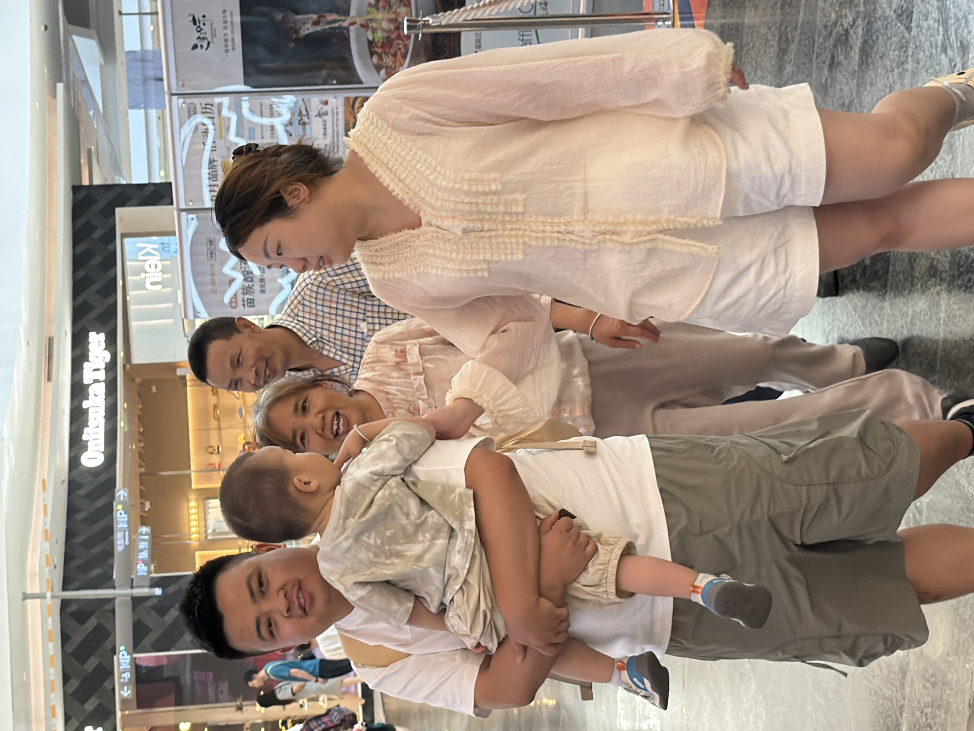 In Yuzhong District, China, a family is walking through an indoor shopping mall. A man, positioned on the left, carries a young child in his arms. To his right, a woman with dark hair pulled back smiles. Behind them, a man in a plaid shirt looks forward. On the far right, a woman in a white blouse and shorts walks alongside, engaging in conversation with the man holding the child. The mall environment features storefronts with signage, including "Onitsuka Tiger" and "Klein," and reflective flooring. The lighting suggests it is daytime.