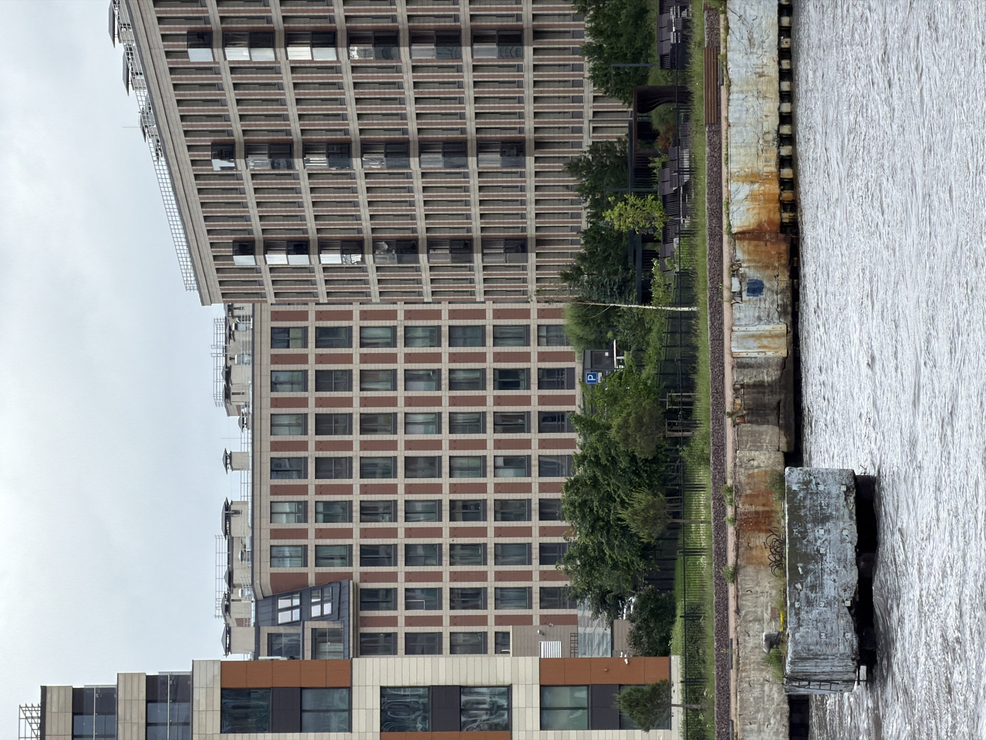 The image shows a view of a waterfront with modern apartment buildings in Saint Petersburg, Russia. In the foreground, a concrete structure, possibly a pier or a breakwater, sits in the choppy water. The structure is weathered and shows signs of rust and damage. Behind the concrete structure, a row of green trees separates the waterfront from a black metal fence. Beyond the fence, the modern apartment buildings are visible. The buildings have a grid-like facade with numerous windows and balconies. The sky is overcast and a pale gray. The apartment buildings are located in the Vasilyevsky Island area, possibly near the Western High-Speed Diameter. There are no people visible in the image.