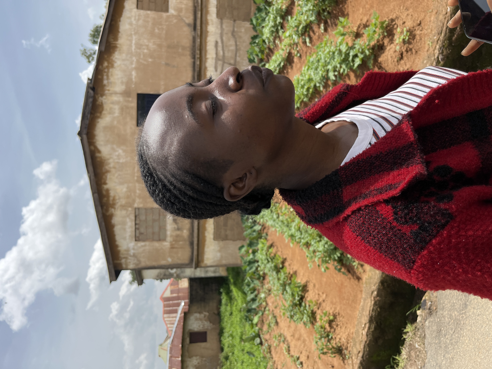 A young Black woman, with her eyes closed and head tilted slightly upwards, is captured in a medium shot from the side in an outdoor, rural setting. She wears a white t-shirt with thin red and black stripes, layered under a red and black checkered knitted jacket. Her hair is styled in cornrows that are pulled back into a bun. The background features a few low-rise buildings with weathered, tan-colored walls and corrugated metal roofs. In the middle ground, rows of green plants grow in rich, reddish-brown soil, suggesting a garden or farm. The sky above is partly cloudy with patches of blue. The overall impression is one of a serene moment in a developing or rural area, possibly in Bukuru, Nigeria.