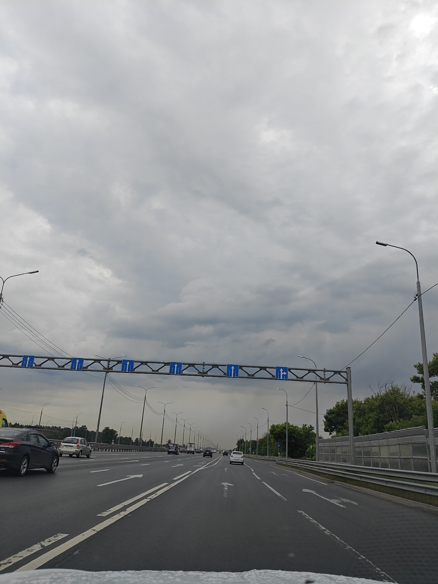 This is a full-shot, eye-level, outdoor, daytime, vertical photograph taken from a moving vehicle on a multi-lane highway with a cloudy sky. The highway is wet, suggesting recent rain, and features white lane markings and arrows indicating traffic flow. Overhead, a large blue and white gantry displays traffic signs with arrows indicating lane directions. Several cars and a truck are visible on the road, all traveling in the same direction. On the right side of the highway, there is a sound barrier and trees. The scene is set in Pushkino, Russia, as indicated by the context.