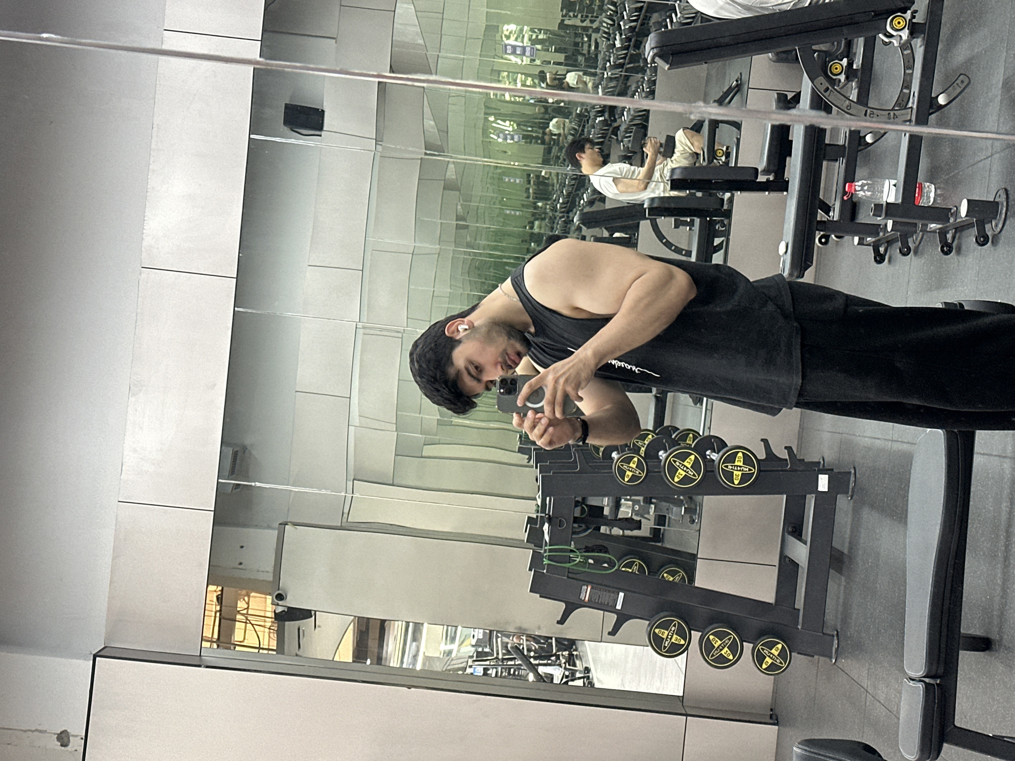 The image depicts a man taking a mirror selfie in a gym. The man, wearing a black tank top and black pants, is positioned facing a wall of mirrors, holding a smartphone with a MagSafe ring to capture his reflection. A white wireless earbud is visible in his left ear. To his right, a black two-tier dumbbell rack displays various dumbbells, some clearly marked with "30" and "MULTI-THI", likely indicating weight. Green fitness bands are also hanging on the rack. A black weight bench is located in the foreground.
Through the multiple reflections on the mirrored wall, another man can be seen in the background, wearing a white t-shirt and light-colored shorts, sitting on a different weight bench. Additional gym equipment, including exercise machines and a red water bottle, are partially visible in the reflections. The lighting suggests it is daytime. A partial brand logo, "supreme," is visible on the main subject's tank top. The setting is clearly a modern fitness center, characterized by its mirrored walls and various workout equipment.