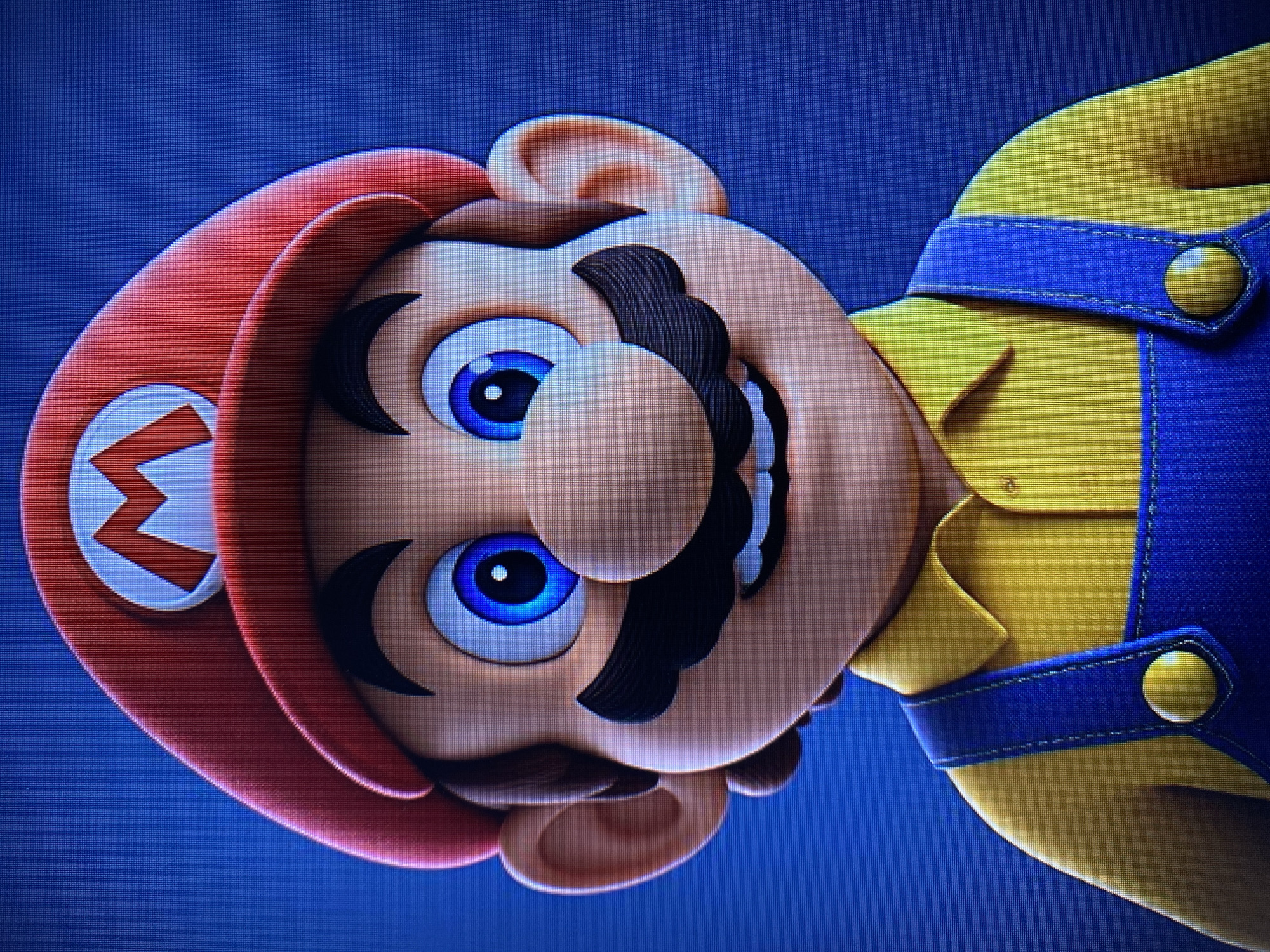 The image is a close-up, head-and-shoulders portrait of a 3D animated character resembling Mario from the Super Mario video game franchise. The character is wearing his iconic red cap with a white circle and a red "M" logo, a yellow collared shirt, and blue overalls with yellow buttons on the straps. He has bright blue eyes, a prominent nose, a bushy black mustache, and a slight smile. The background is a solid, dark blue. There is no indication of any location in Yerevan, Armenia, or any urban environment.