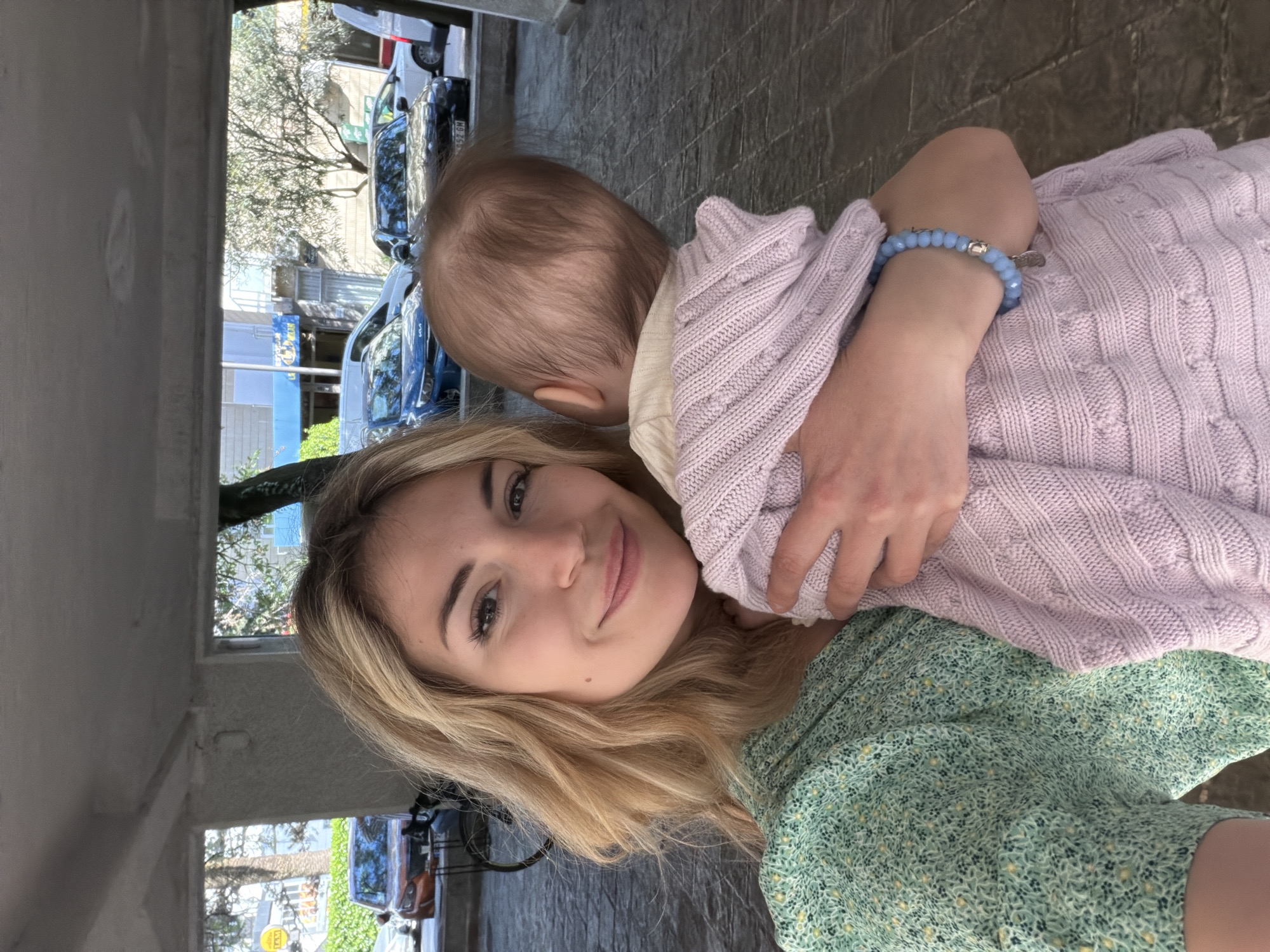 A close-up, chest-up selfie captures a woman holding a baby outdoors in Budva, Montenegro, during daylight hours. The woman, with long blonde hair, smiles warmly at the camera. She is wearing a light green dress with a small, intricate floral pattern. On her left wrist, a light blue beaded bracelet with a silver charm is visible.
She is holding a baby, whose back is towards the camera. The baby has light brown hair and is dressed in a textured, light purple knitted sweater over a light yellow undergarment. The woman's arm is visible around the baby, suggesting a gentle embrace.
The background reveals an urban street scene, possibly viewed from under a covered walkway or awning, indicated by the partial view of a light-colored ceiling overhead. Several cars are parked on the street, including blue and white vehicles. A yellow "TAXI" sign is clearly visible on a building across the street. Other buildings and trees with sparse foliage and green leaves are also in the background, along with a glimpse of a bright sky in reflections. Part of a car license plate, "HV 926M," is also discernible. The setting suggests a casual, everyday moment in a town environment.