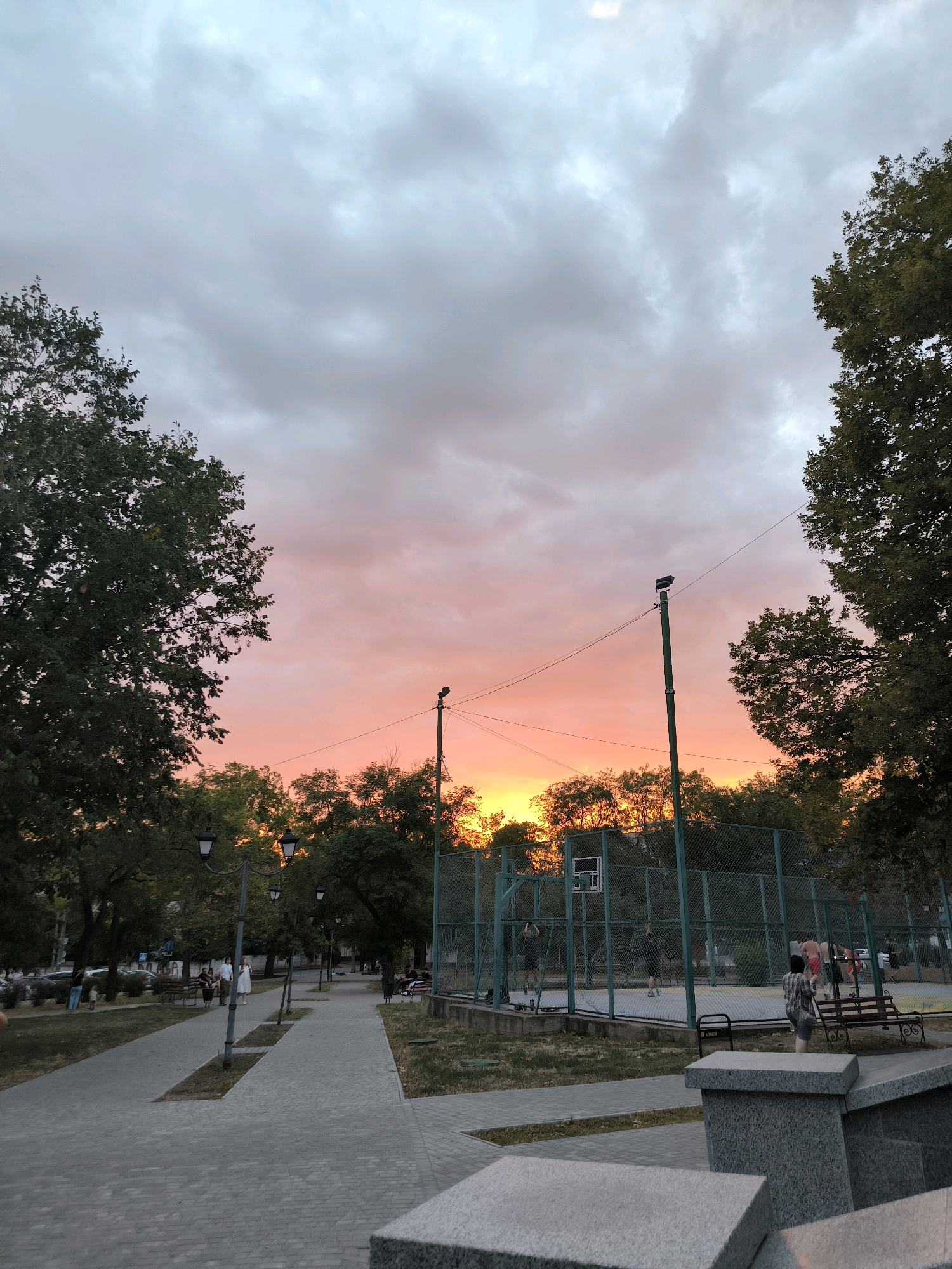This image depicts a sunset over a park in Mykolaiv, Ukraine. The sky is filled with dramatic clouds painted in hues of pink, orange, and gray. In the foreground, a paved walkway winds through a park with lush green trees lining its sides. Several lampposts are visible along the walkway, and benches are scattered throughout the park.
To the right of the walkway, a basketball court is enclosed by a green metal fence. Several people are visible on or around the court, some appearing to be playing basketball, while others are observing. The overall atmosphere of the scene is serene and peaceful, with the vibrant colors of the sunset creating a beautiful backdrop.
The image captures a moment of leisure and community within the urban landscape of Mykolaiv. The presence of the basketball court suggests a recreational area, while the park setting provides a natural respite within the city. The details within the image, such as the paved walkways and lampposts, indicate a well-maintained public space.