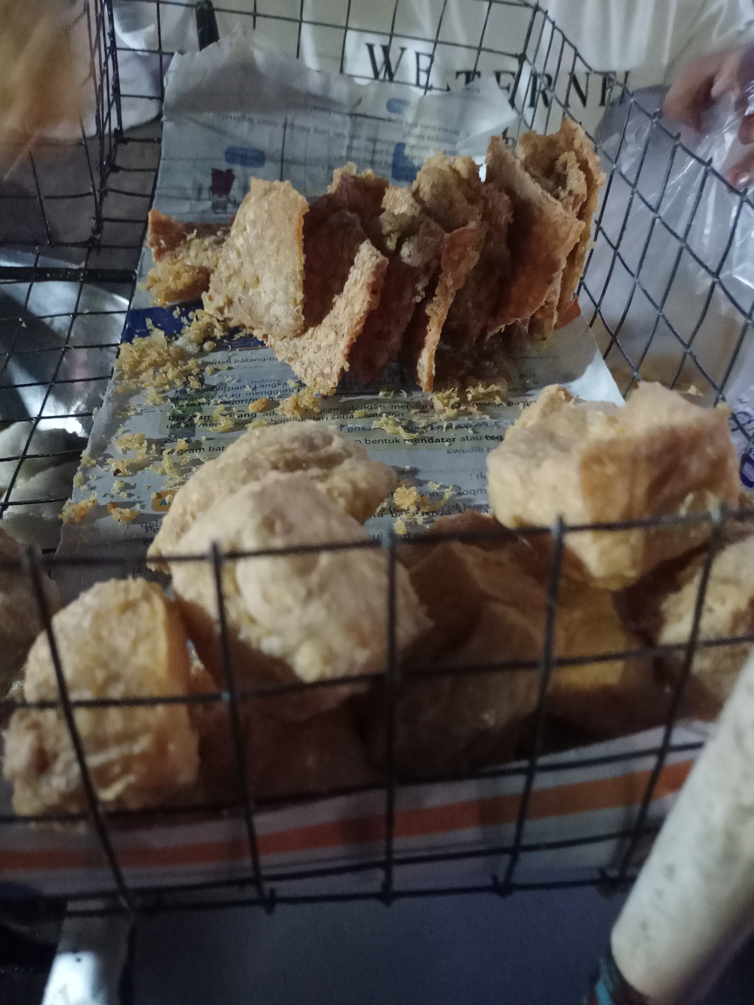 This image captures a close-up view of fried tofu being displayed and sold in a wire basket. The tofu pieces vary in shape and size, with some appearing to be whole cubes and others cut into thinner, more irregular pieces, all coated in a light brown batter. They are arranged on top of what appears to be newspaper, which is partially visible beneath the food. The basket itself is made of black wire mesh, and it is placed in an outdoor setting, likely a street food stall. In the background, a white t-shirt with the partially visible text "WETERN" can be seen, along with a hand holding a clear plastic bag, suggesting a customer interaction. The overall lighting suggests it might be evening or nighttime. The scene is located in the city of Karawang, Indonesia, a region known for its diverse street food culture.