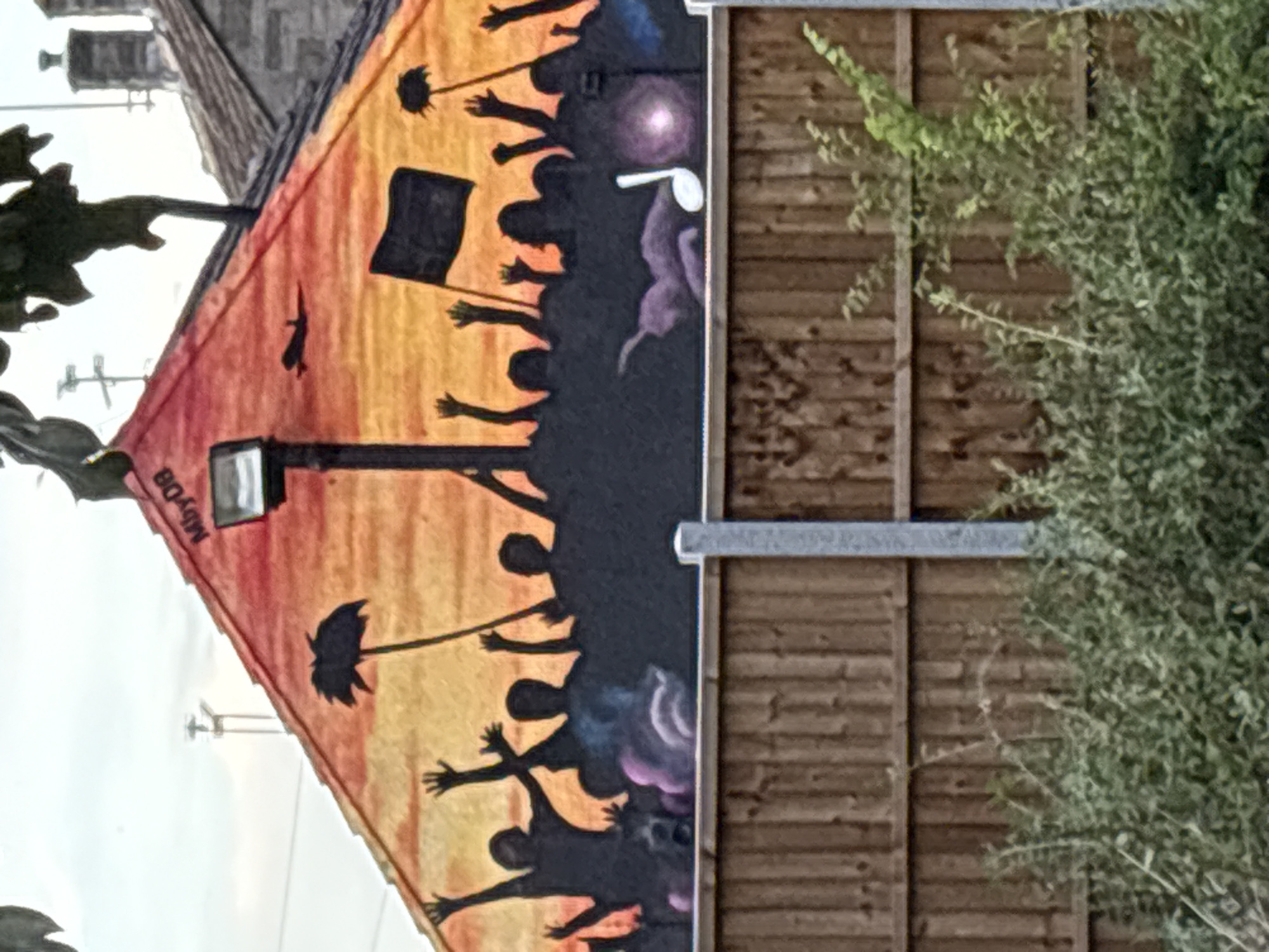 The image shows a mural on the side of a building, depicting a crowd of silhouetted figures with their arms raised. Some figures are holding umbrellas, while one holds a black flag. The mural is painted in warm colors, with a gradient from orange to yellow at the bottom, transitioning to a darker, purplish hue at the top. A light fixture is visible on the wall, extending into the mural. The mural is adjacent to a wooden fence with a metal railing. To the left of the mural, there is a partial view of other buildings, including what appears to be a brick structure, and some overhead wires against a bright, overcast sky, suggesting it is daytime. The overall impression is one of a protest or a celebratory gathering. There is visible text on the mural which reads "MByD8".