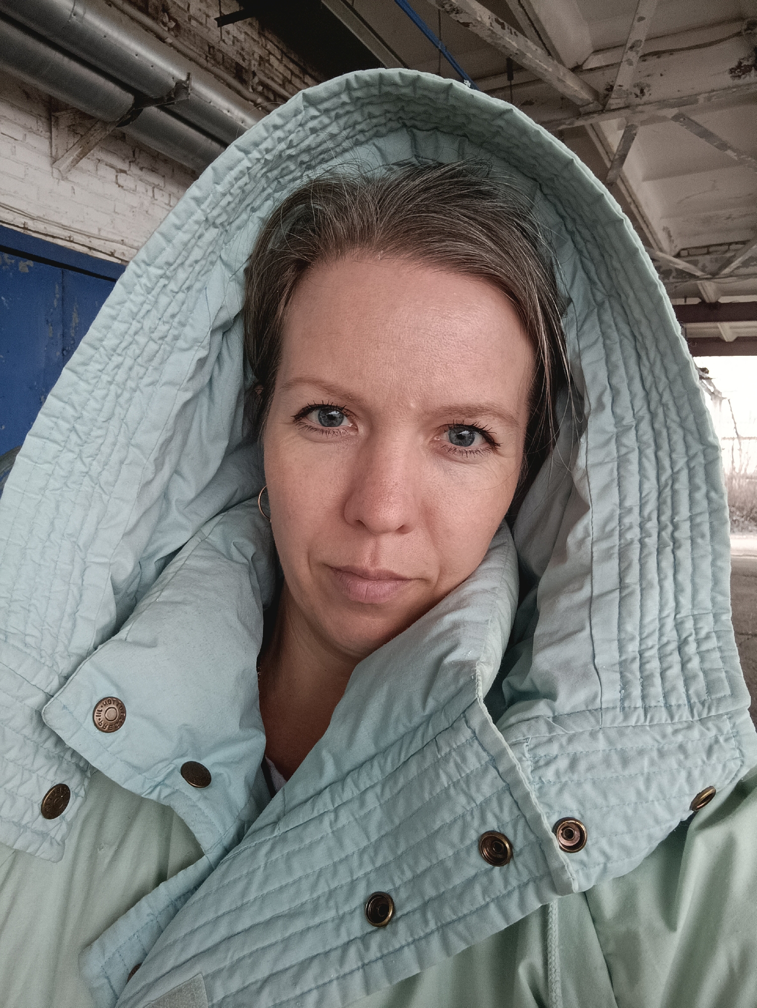 A medium, eye-level shot depicts a woman wearing a light blue, quilted hooded jacket. Her eyes are blue, and she is looking directly at the camera with a neutral expression. Her hair is graying. Gold hoop earrings are visible.
The background is a somewhat industrial setting, suggesting an outdoor but covered location. A blue metal door is partially visible in the upper left corner. In the upper center and right, there's a view of a concrete ceiling structure with exposed beams. The photograph appears to have been taken outside in Chaikovsky, Russia.