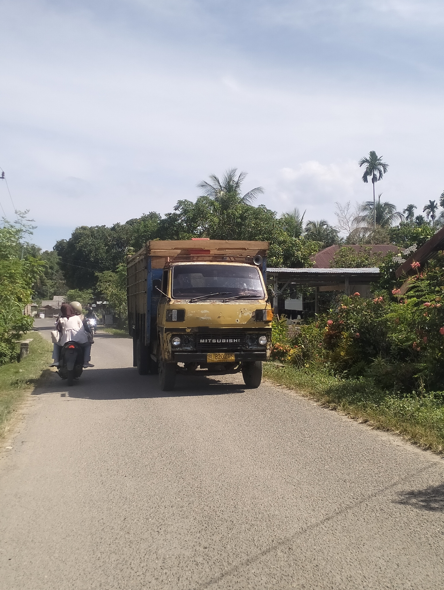 A yellow Mitsubishi truck is traveling down a paved road in Meunasahbuket, Indonesia. The truck is loaded with wooden planks and is being passed by a motorcycle carrying two people. The motorcycle riders are wearing helmets. The road is flanked by lush green vegetation and trees, with several houses visible in the background. The sky is partly cloudy.
