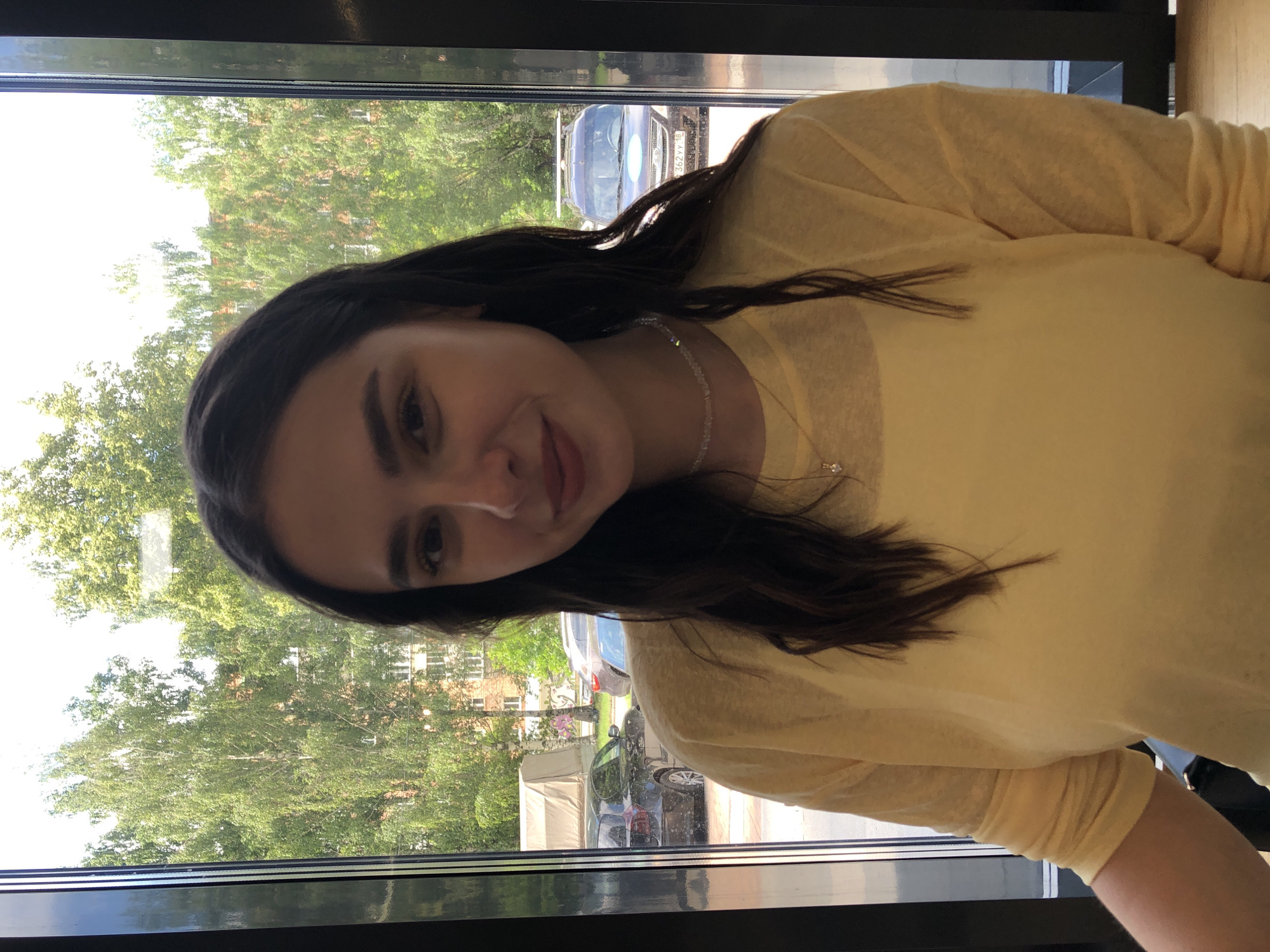A young woman with long, dark hair is shown from the chest up, turned towards the camera. She is wearing a light yellow, long-sleeved shirt and a delicate necklace with a small pendant. Her expression is neutral, with a slight smile. The background suggests an indoor setting with a large window. Through the window, trees with green foliage and some buildings are visible, indicating an outdoor scene. Several cars are parked outside, one with a license plate that appears to have Cyrillic characters. The lighting suggests it is daytime, likely sunny, given the brightness and shadows. The woman is the sole subject of the photograph.