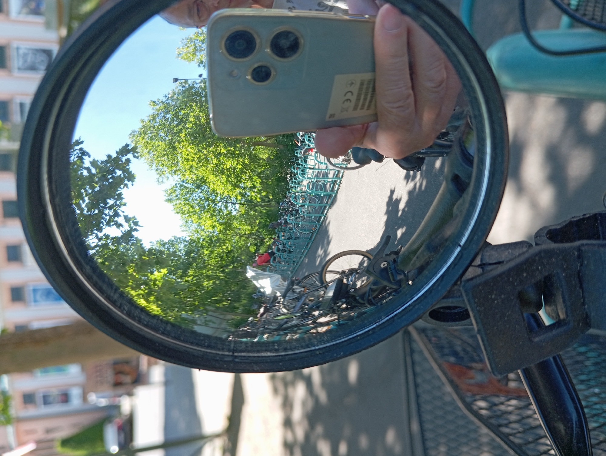 The image is a selfie taken in a bicycle mirror, reflecting a street scene in Baden, Austria. In the foreground, the mirror's rim is visible, along with parts of the bicycle's handlebars and a basket. The reflection in the mirror shows a row of parked bicycles on a paved surface, with a bright blue bicycle rack. Trees with green foliage are prominent in the background, with a clear blue sky above. In the far distance, a building with an ornate facade and windows is partially visible. A person's hand holding a smartphone, used to take the selfie, is also reflected in the mirror, along with a portion of the person's face. The overall setting appears to be a sunny day in an urban or park-like area.