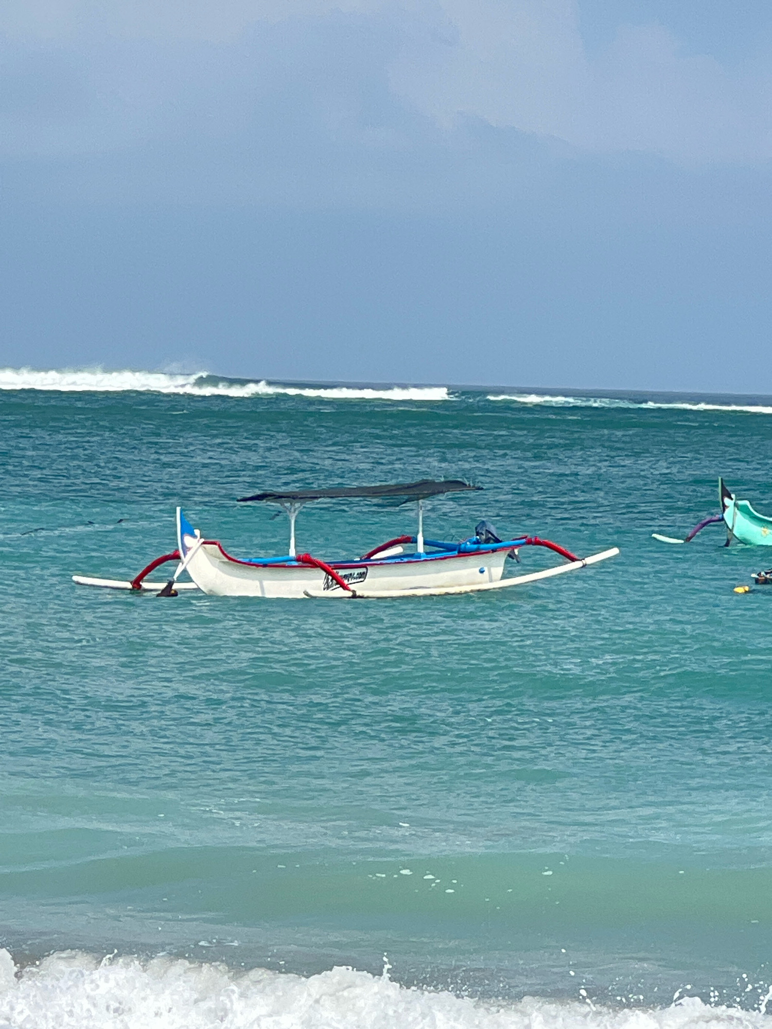 A traditional Indonesian fishing boat, known as a jukung, floats on the turquoise waters off the coast of Kuta, Indonesia. The boat, primarily white with red and blue accents, features a canopy providing shade. In the background, a line of waves breaks on the shore, indicating a moderately active sea. Another, smaller boat is visible to the right. The sky is a pale blue, with some clouds present. The scene depicts a calm, coastal environment.