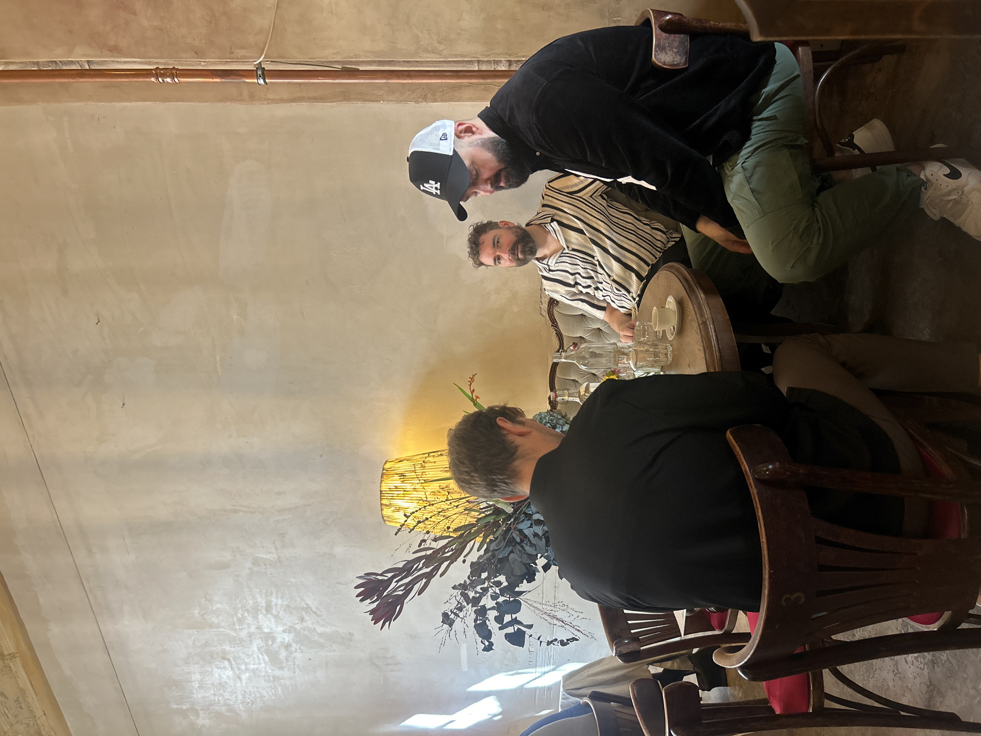 Three men are seated at a round wooden table in what appears to be a cafe in Berlin, Germany. The man closest to the foreground is wearing a black jacket and green pants, with a black and white baseball cap. He is looking down at the table. The man in the middle is wearing a striped shirt and a beard, and is looking towards the man in the cap. The man furthest from the foreground is wearing a black jacket and is mostly obscured from view, facing away from the camera. On the table, there are drinks, glassware, and a small white teacup and saucer. Behind the table, there is a decorative lamp with a patterned shade and a floral arrangement. The background is a textured, light-colored wall with some visible piping. The overall impression is that of a casual meeting or gathering.