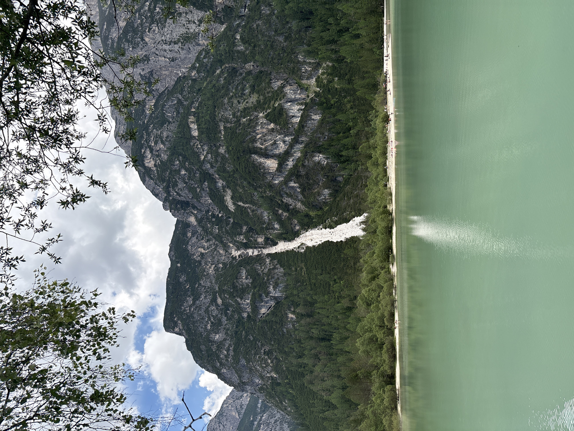 This is a vertical, eye-level, outdoor, medium shot of a serene, aquamarine lake in front of a large, tree-covered mountain range. The lake fills the bottom two-thirds of the frame, with the shoreline and mountain range above. The water is calm with gentle ripples, and a white sandy beach lined with trees and scattered people is visible along the shoreline. The mountain range is steep and rocky, with a significant white scar running down its center, possibly a rockslide or avalanche. The sky above is partly cloudy with patches of blue. The foreground is dominated by the lake, while the midground features the beach and trees, and the background is the imposing mountain. The scene appears to be a natural landscape, likely a park or recreational area.