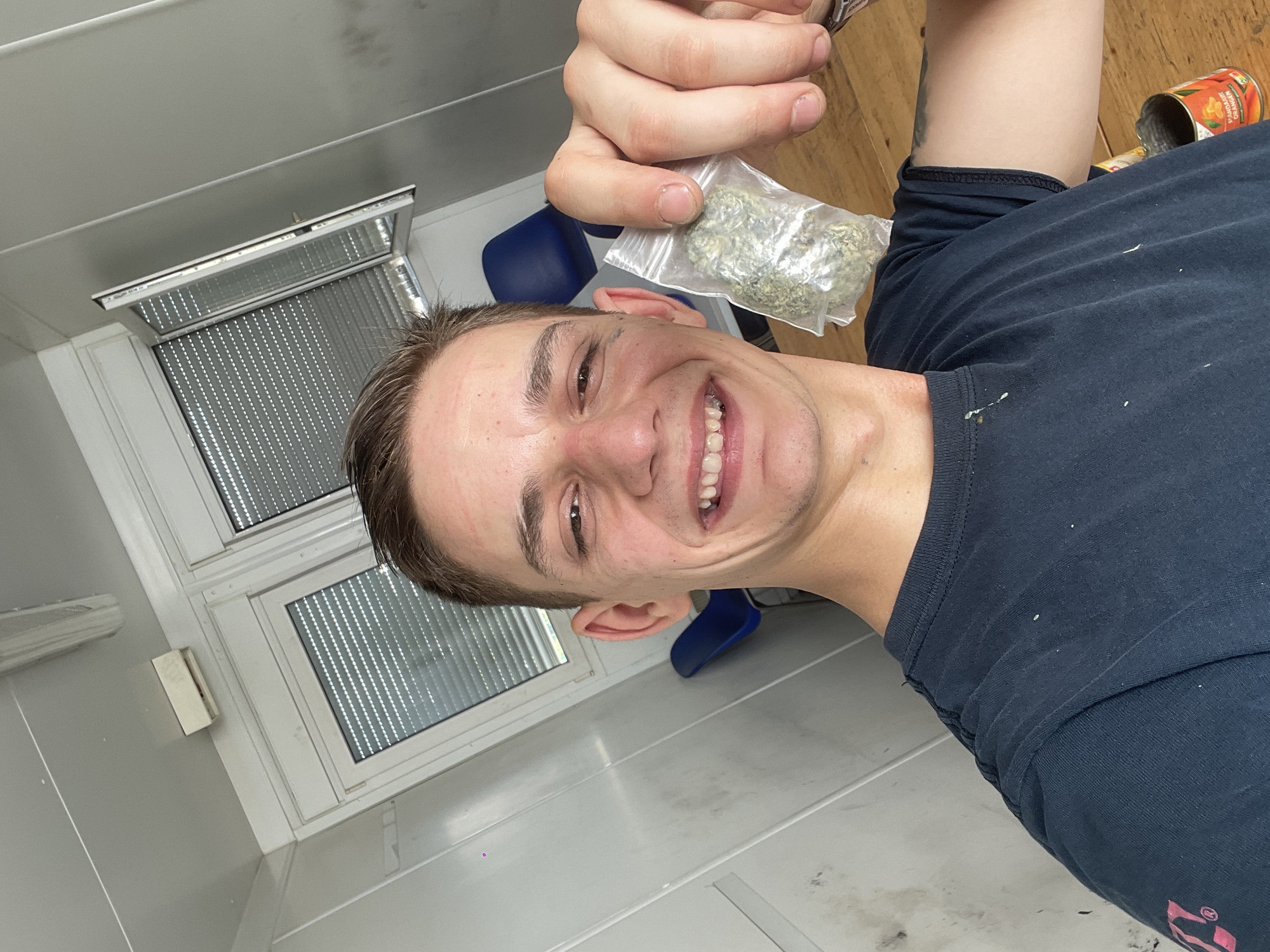 A young adult male is posing for a selfie in a room with a window and metal blinds. The room appears to be a basic, prefabricated structure with white walls and gray flooring. The male is wearing a dark-colored t-shirt and is holding a small plastic bag containing green leafy material. There is a blue chair partially visible in the background, as well as an open can on the right side of the frame. Location is Unknown, Unknown.