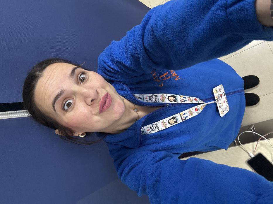 A woman takes a selfie from an elevated perspective, looking directly at the camera with wide eyes and pursed lips. She has dark hair pulled back, visible freckles, and wears a royal blue fleece jacket with a partially open zipper. A white lanyard featuring colorful cartoon characters and text, including "KEEP CALM I'M A NURSE" and "DANCE IT OUT," hangs around her neck, holding an identification badge. Her jacket is embroidered on the right side with orange text, "ILAYAT H Meknès," confirming the location as Meknes, Morocco, likely within a healthcare institution. She also wears a subtle necklace with a small charm and gold-colored hoop earrings. Her feet, clad in black flat shoes, are visible at the bottom of the frame. To the lower left, a black smartphone with a charging cable rests on a light-colored surface. The background consists primarily of a plain dark blue wall or partition, with a vertical metallic strip visible on the right side.