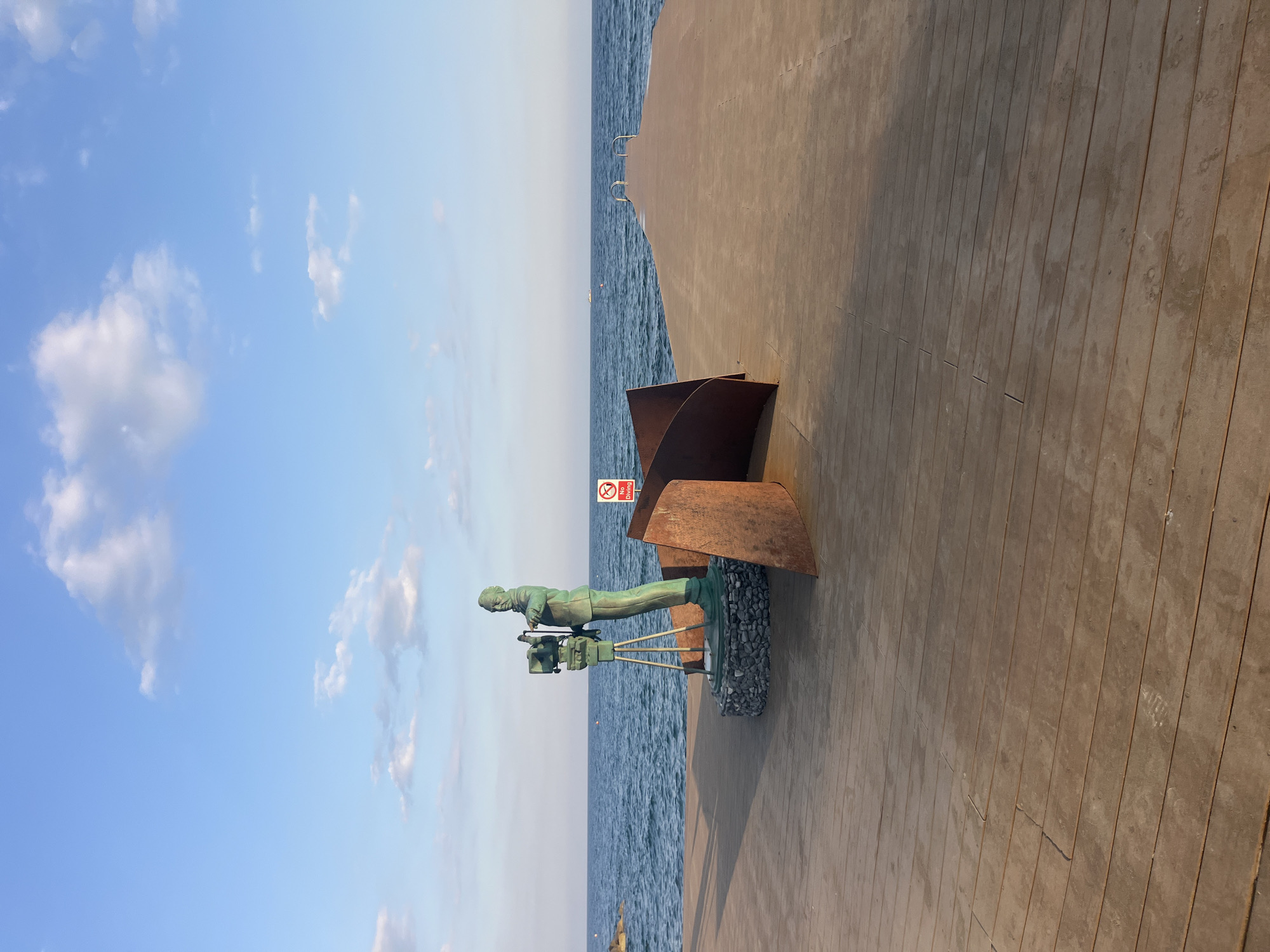 The image depicts a sculpture of a person operating a vintage film camera, located on a wooden pier extending into the sea. The sculpture, made of a greenish-bronze material, stands on a circular base of dark stones. To the right of the sculpture are two abstract, rusted metal panels forming a curved shape. A "No Diving" sign is visible behind the sculpture. The wooden pier leads towards the sea, with a gentle curve and metal railings at its end. The sea is a deep blue with moderate waves, and the sky above is a clear blue with scattered white clouds. The overall scene suggests an outdoor public space, likely a waterfront promenade or park in the Agios Tychona Community, Cyprus, given the presence of the sea and what appears to be a resort-style setting. The lighting indicates it is either early morning or late afternoon, casting shadows across the pier. There are no people actively interacting in the scene, only the static representation of a cinematographer and the environment itself.