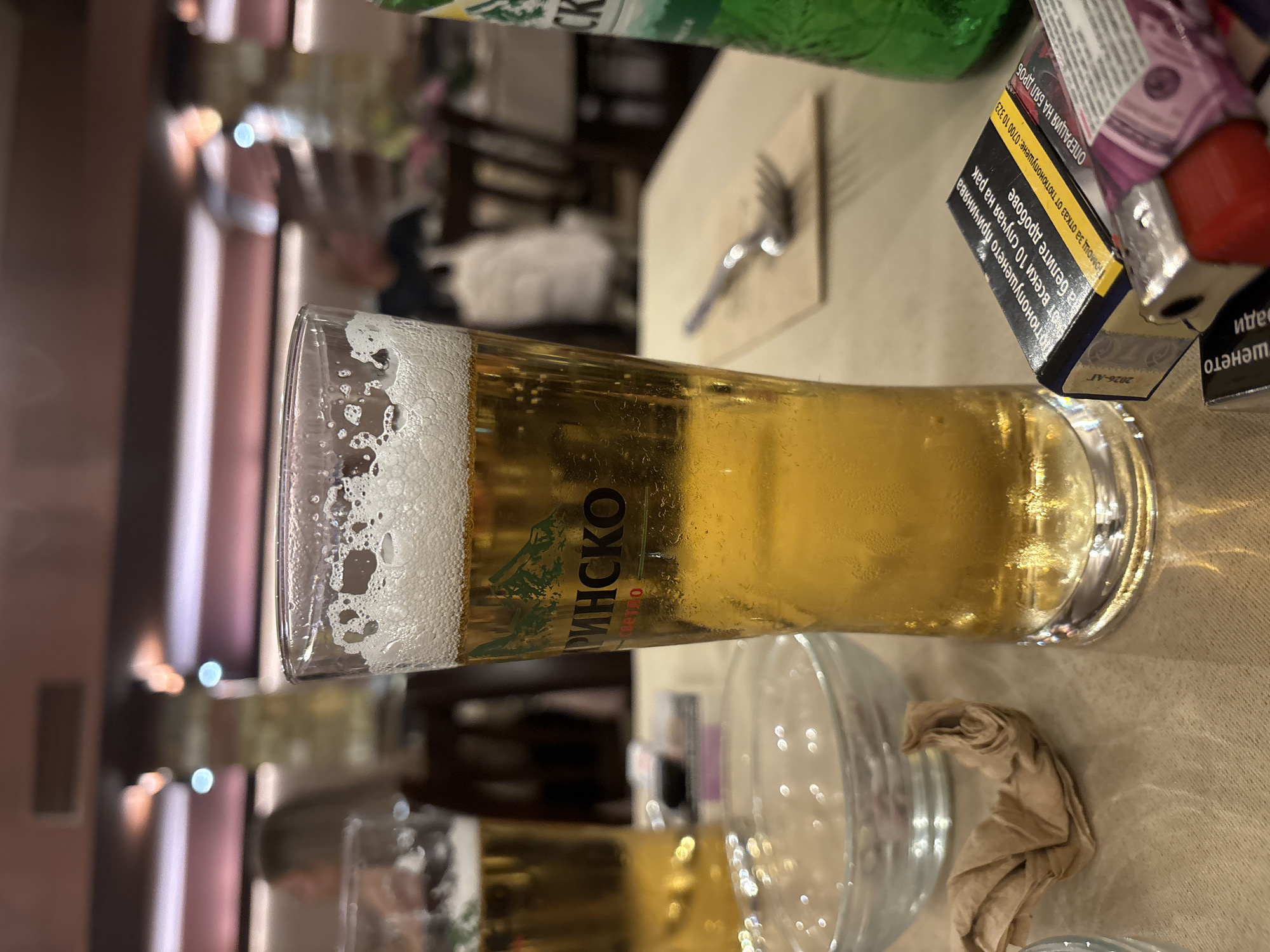 The image shows a glass of "Pirinsko" beer on a table, likely in a restaurant setting, given the presence of a fork, napkins, and a blurred background suggesting other tables and patrons. There's also a pack of cigarettes with text in Bulgarian, a lighter, and a half-empty glass nearby. Due to limited visual information, pinpointing the exact location in Pleven, Bulgaria is impossible. However, the details like the beer brand and cigarette packaging suggest the setting is within Bulgaria.