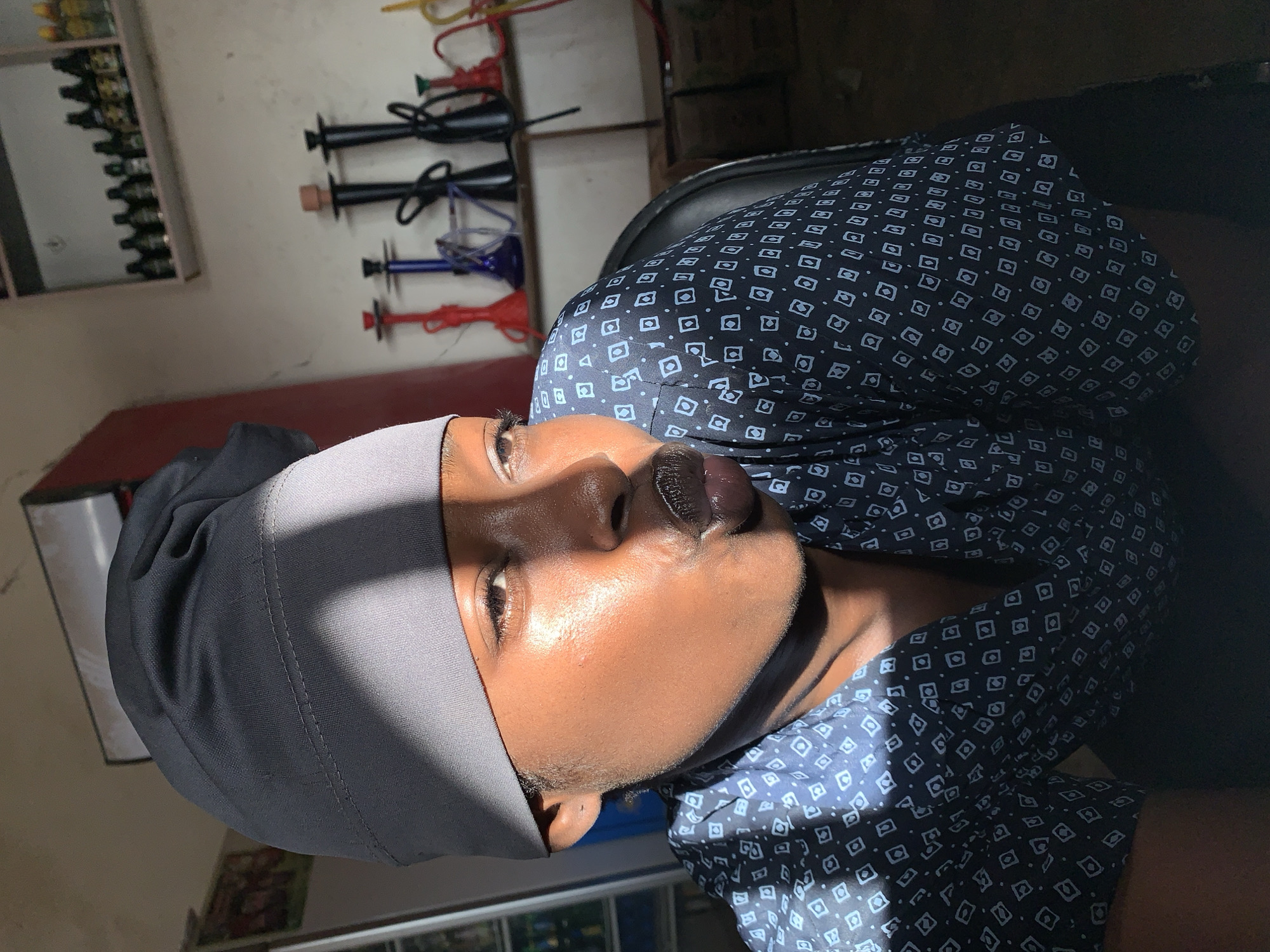 A person with dark skin, wearing a dark head covering and a blue shirt with a geometric pattern, is positioned in front of shelves displaying bottles and hookah equipment. The setting appears to be indoors.