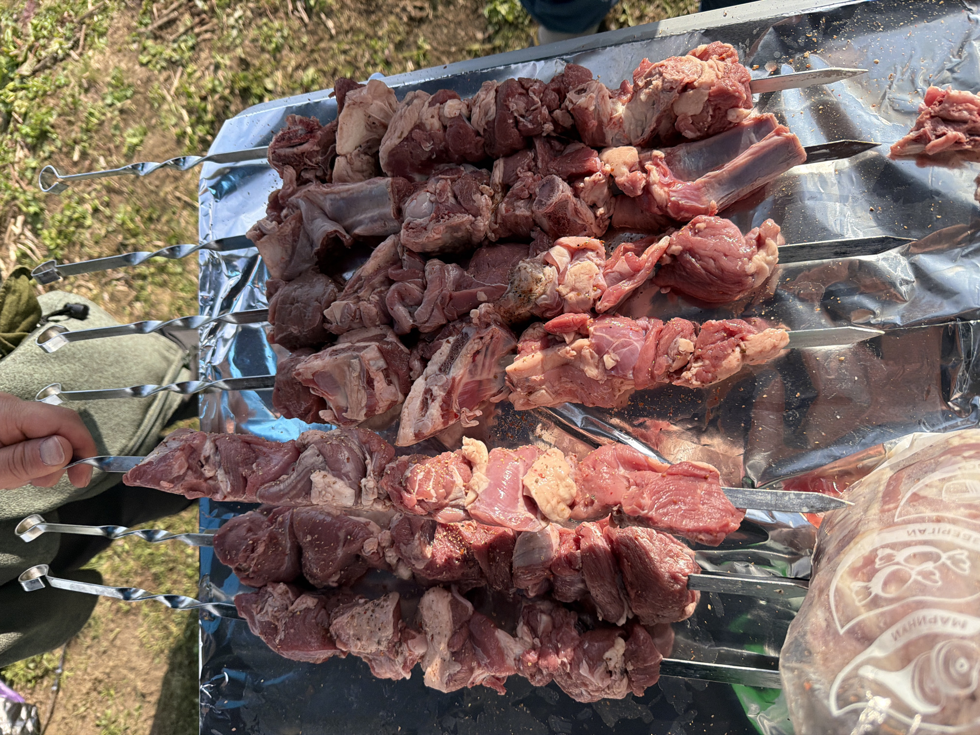 In an outdoor setting, likely in or near Mestia, Georgia, a person is preparing raw meat skewers for cooking. The skewers, made of metal with twisted shafts, are loaded with cubed pieces of red meat, some of which appear to be lamb or mutton, interspersed with fatty sections and bone fragments. The meat is seasoned with visible spices. These skewers are laid out on a surface covered with reflective foil. In the foreground, a portion of a plastic package containing raw meat is visible, with Cyrillic lettering on it. The background consists of dry, grassy ground and some vegetation, suggesting a rural or park-like environment. The lighting indicates it is daytime, possibly late afternoon, with shadows cast by the skewers and meat.