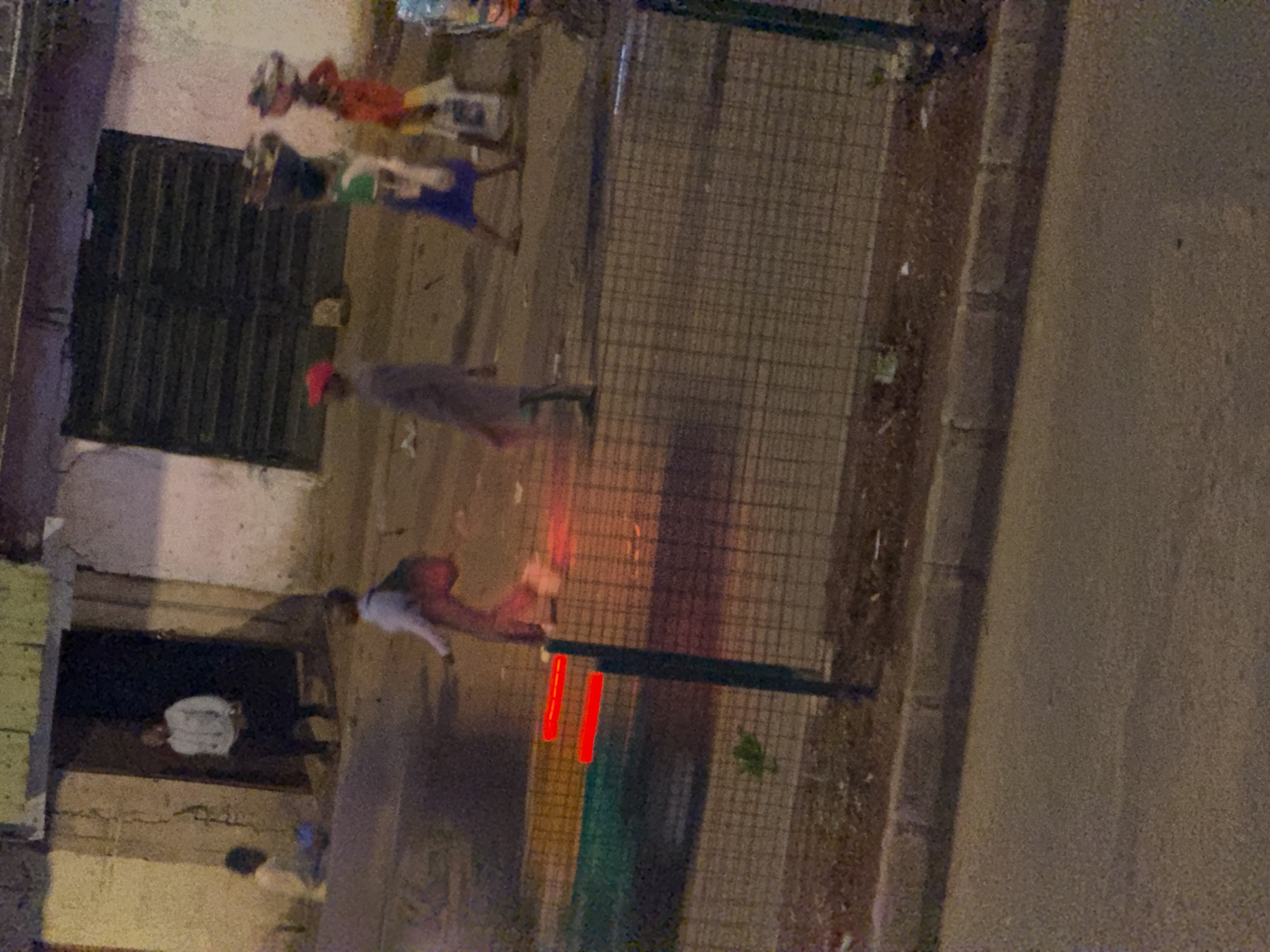 The image depicts a street scene in Bukuru, Nigeria, at dusk or night. In the foreground, a sidewalk is separated from a road by a curb. A wire fence runs along the edge of the sidewalk. Two red horizontal lines, possibly part of a traffic light or other illuminated signage, are visible on a dark post in the foreground.
Further back, several individuals are visible. On the left, a person in a light-colored top and dark pants stands in a doorway. To their left, a child in a light-colored garment is seated on the ground. In the middle of the street, a person wearing a grey outfit and a red cap walks from right to left. Behind this individual, three people are walking from right to left, each carrying baskets on their heads. The person in the middle of this group wears a green top and blue shorts, while the person to their right wears a red top and white bottoms.
The background features a building with a dark, open doorway and shuttered windows. The ground appears to be paved. The lighting suggests artificial sources, contributing to the low-light conditions of the scene.