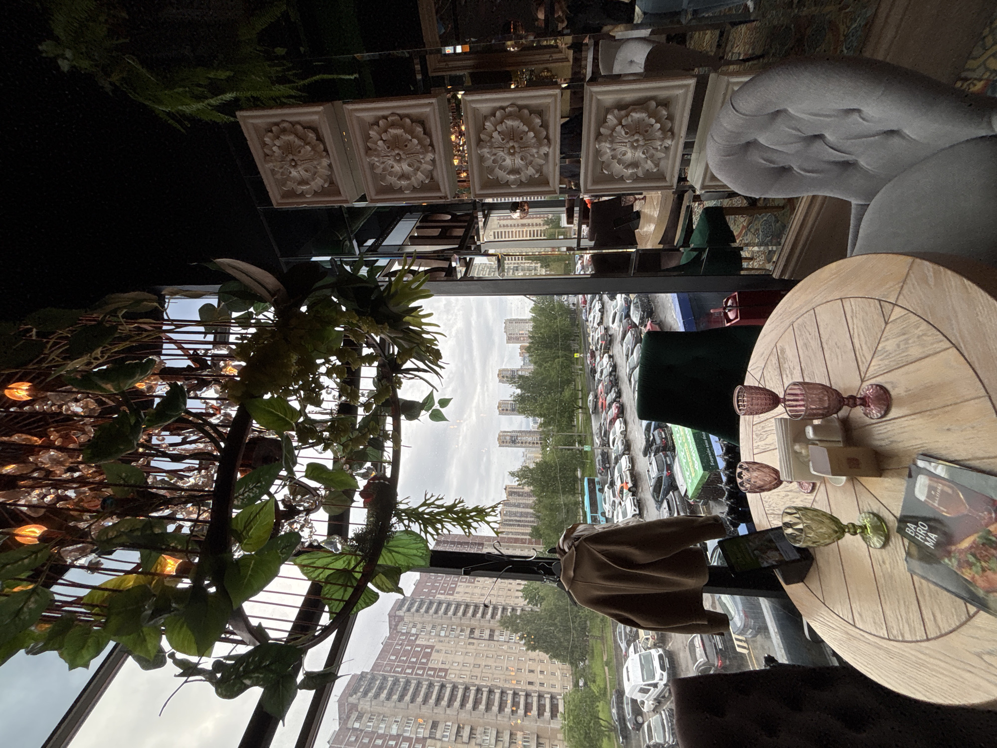 The image depicts a scene from what appears to be a restaurant interior in Saint Petersburg, Russia, as evidenced by the architecture seen through the window.
**Environment:** The restaurant has ornate interior decorations, including ceiling embellishments with floral motifs and decorative plants. The view through the window reveals a cityscape with residential buildings and a large parking area filled with cars. The sky is overcast.
**Objects:** A round wooden table is the focal point, set with glassware (three goblets, two of which appear pink and one green), napkins, and a menu. There is a jacket draped over a chair near the window. A sofa is visible in the periphery.
**Subjects/People:** No people are clearly visible, but the presence of set table and jacket suggests occupancy. The restaurant is near residential buildings.
**Actions/Interactions:** The scene shows a prepared table setting, indicative of anticipated or recent dining activity.