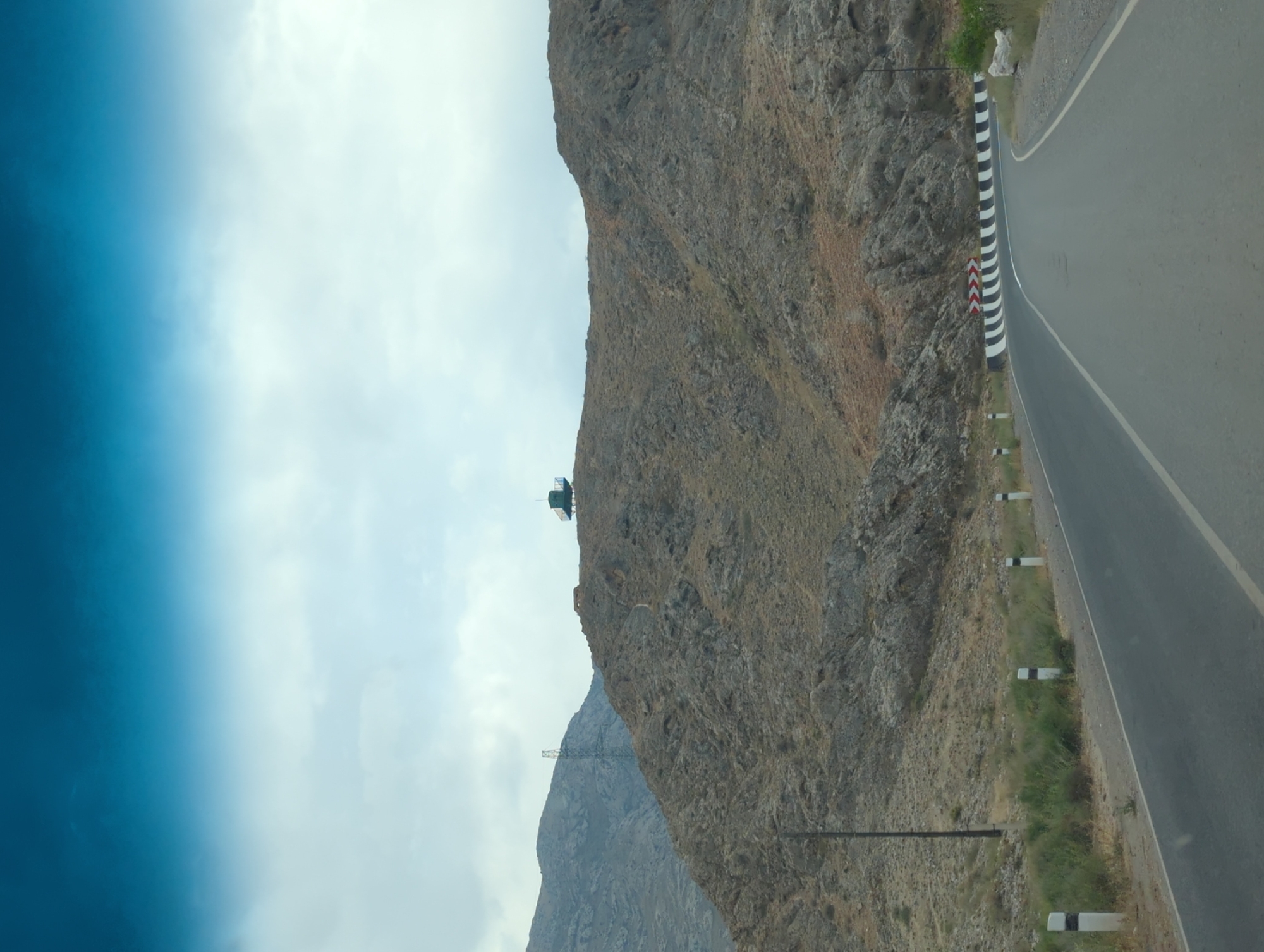 The image depicts a view from a vehicle traveling on a paved road through a mountainous, arid landscape. The road curves to the right, bordered by white posts with black reflective bands. To the left of the road, sparse vegetation grows at the base of rocky hills. In the distance, a tall electrical pylon stands on a higher mountain slope. On a prominent ridge in the center of the image, a small, greenish-blue structure with a raised platform and railings is visible. The sky above is overcast with diffused light. The scene appears to be located near the city of Kök-Tash, Kyrgyzstan, given the topography and style of infrastructure.