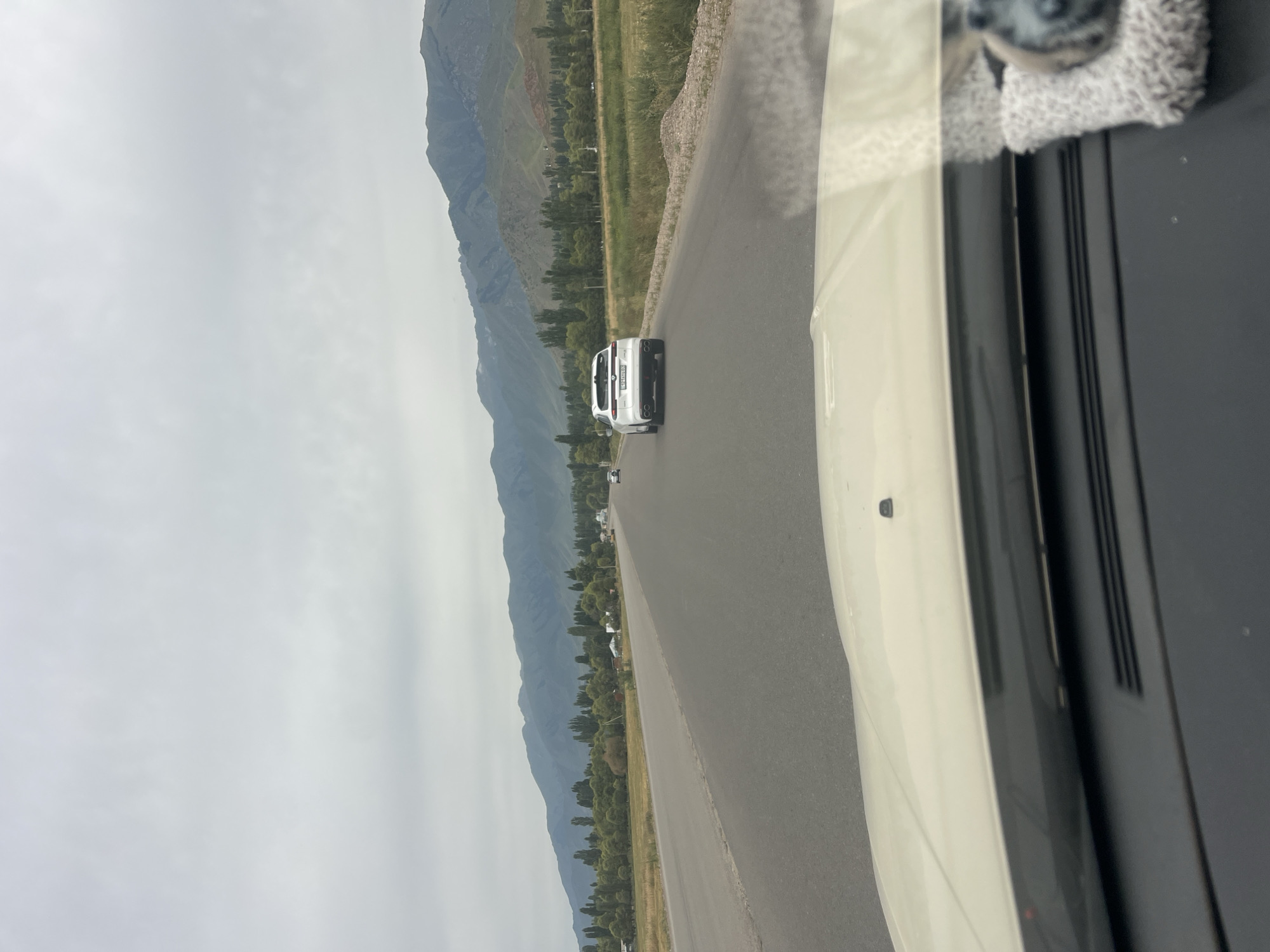 The image shows a view from inside a vehicle, looking forward along a two-lane asphalt road. The road stretches into the distance, with mountains in the background. A white SUV is visible on the road ahead, moving in the same direction. The landscape includes fields and trees on either side of the road, with some buildings scattered amongst the trees. The sky is overcast. The specific location is identified as being in the city of Balbay, Kyrgyzstan.