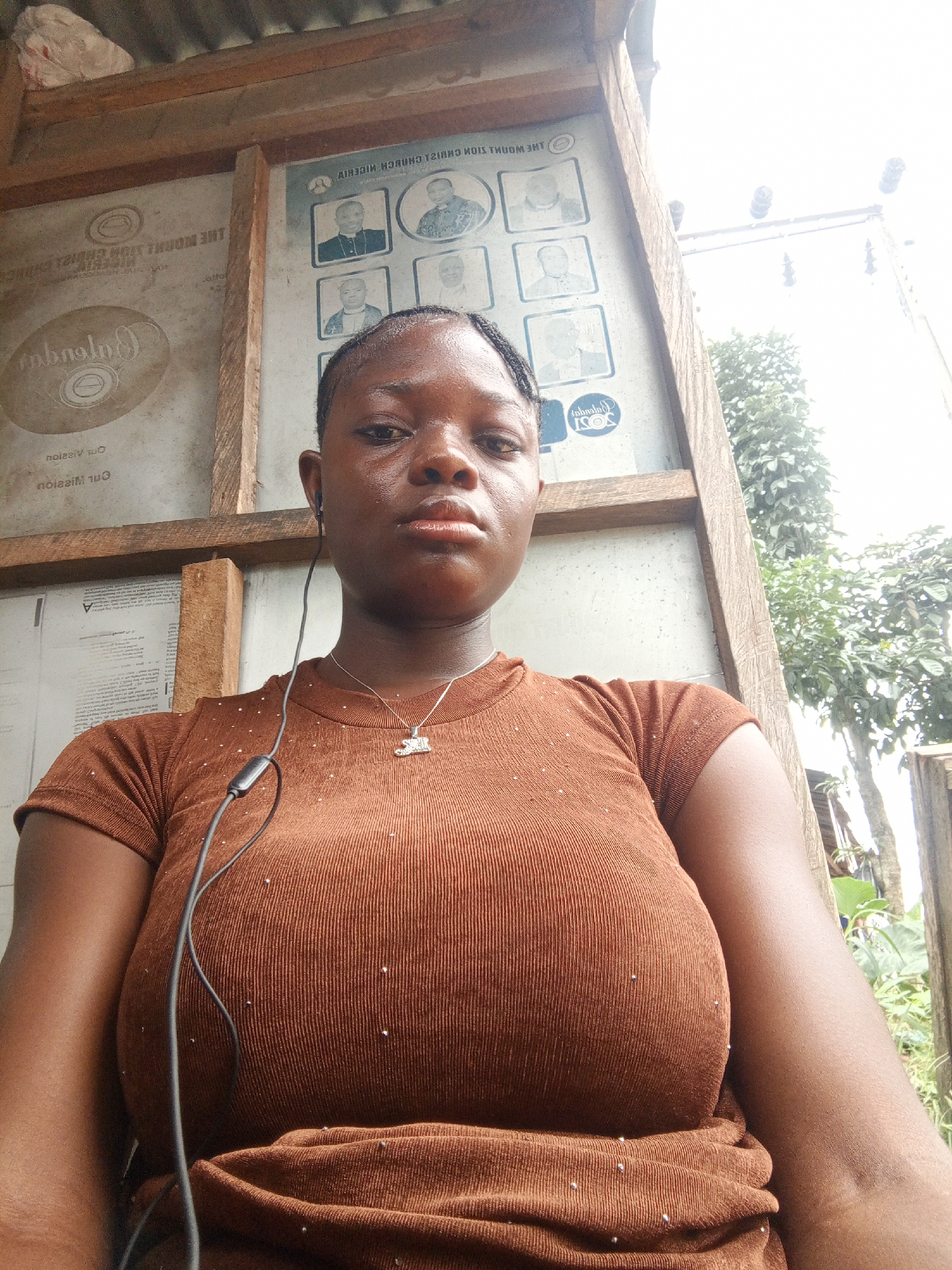 A woman is standing outdoors in Mbribit, Nigeria. She is wearing a brown shirt and a necklace, with earbuds in her ear. Behind her is a wooden structure, possibly a bus stop or shelter, with signage that includes photos of people and text in both English and potentially another language. To the right of the structure, there are trees and glimpses of the outside environment. The area appears to be relatively simple and perhaps rural, though the presence of electrical wires suggests some infrastructure.