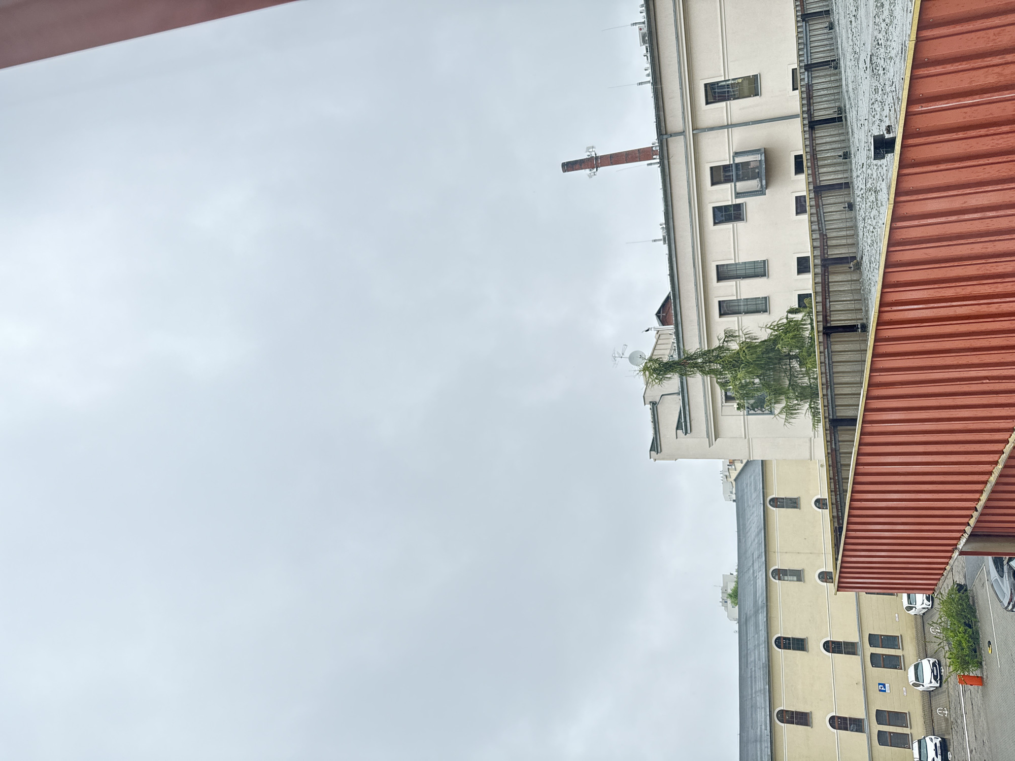 The image captures a view of buildings under an overcast sky, suggesting a cloudy day. The perspective is from a higher vantage point, looking down and to the side at several structures.
On the right side of the frame, a corrugated red roof is prominent, angling downwards. Adjacent to this is a multi-story building with a light-colored facade and a dark roof. This building features numerous windows, some with arched tops, and several doors on its lower levels. A row of parked cars, mostly white, is visible on the pavement in front of this building. Some of these cars are facing upwards, indicating the image is rotated. A small green bush or tree is planted in front of one of the cars. A blue sign with a white 'P' is visible near the parked cars, likely indicating parking.
Further back and to the left, another taller building is visible, also with a light-colored facade and numerous windows. A brick chimney extends upwards from this building, topped with some antenna-like structures. A tree with wispy green foliage grows near this building, partially obscuring some of its windows and balconies.
The overall scene appears to be an urban or industrial setting, possibly a complex of warehouses or apartment blocks, given the architecture and the presence of a parking lot. The lack of direct sunlight and the gray sky indicate overcast weather. The image orientation is noticeably off, requiring rotation for proper viewing. No people are visible in the frame.