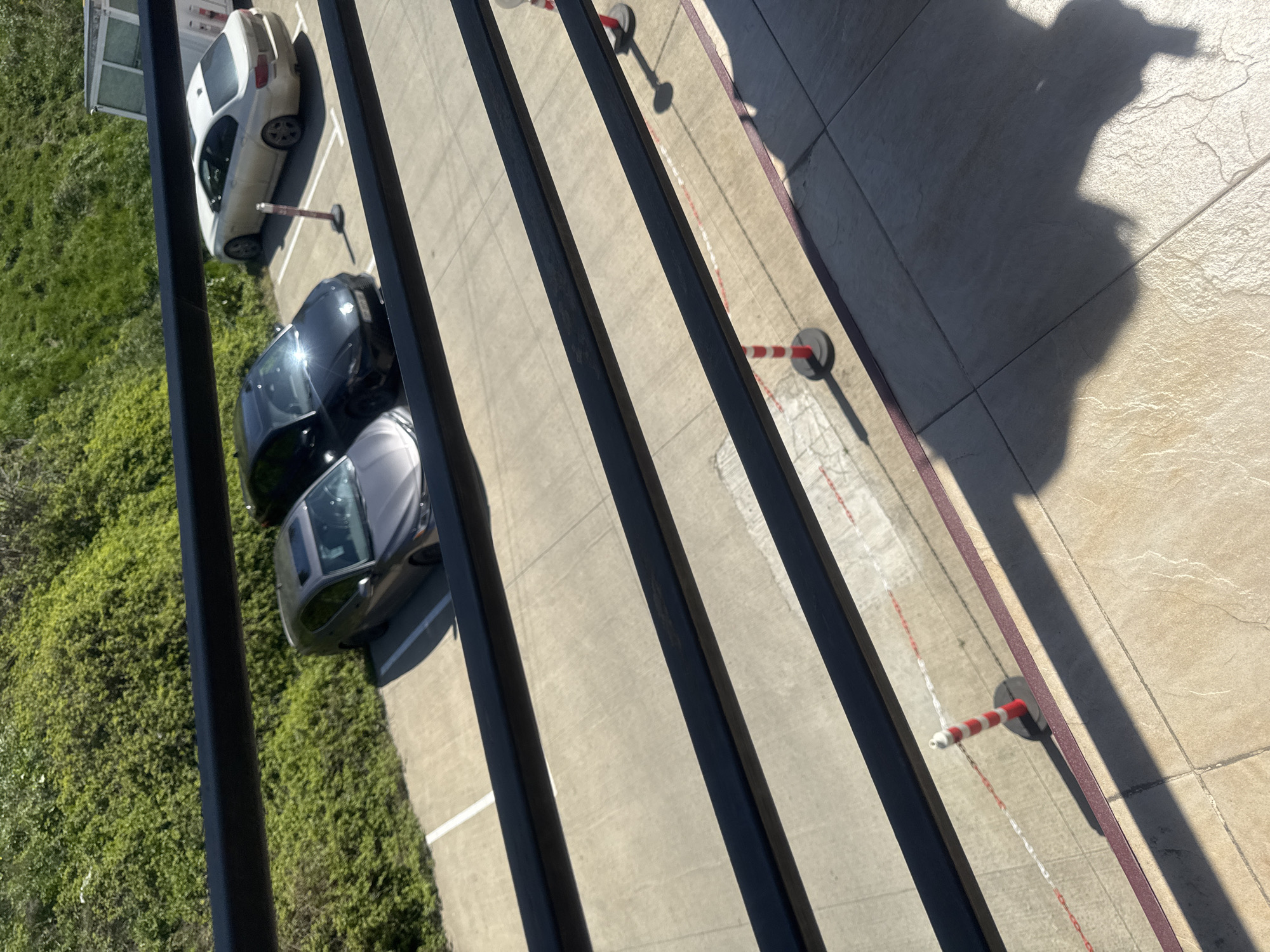 The image, captured from an elevated perspective, depicts a parking area in what appears to be Tbilisi, Georgia. The scene is characterized by three parked cars, one white, one black, and one silver, along with several red and white striped bollards lining the edge of the parking area. Adjacent to the parking spots is a patch of lush greenery. The perspective is from a balcony or high window, with the black railings of the balcony in the foreground. A shadow of a person is visible, most likely cast by the photographer on the tiled ground. The parking surface is concrete, showing signs of wear and use.