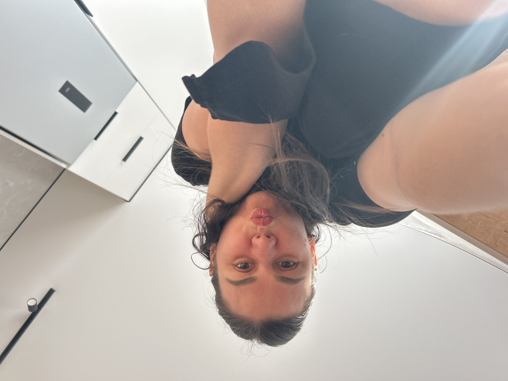 The image is an upside-down, close-up, eye-level shot of a young woman. She is wearing a black t-shirt and has long, dark hair. Her face is turned towards the camera, and she is puckering her lips. Her eyes are open and looking directly at the camera. The background is a brightly lit room with white walls and what appears to be modern cabinetry with dark handles. To the left of the woman, there are two cabinets, one with a light gray surface and a dark rectangular handle, and another with a white surface and two dark rectangular handles. In the lower left corner, there is a black rod with a small white object on it, against a white wall. The overall impression is that the woman is in a domestic setting, possibly a kitchen or bathroom, and is taking a selfie. There is no visible text in the image. The time of day and weather are not discernible. The woman's expression appears playful or flirtatious. The context of Chaikovsky, Russia, does not provide further specific details that can be seen in the image.