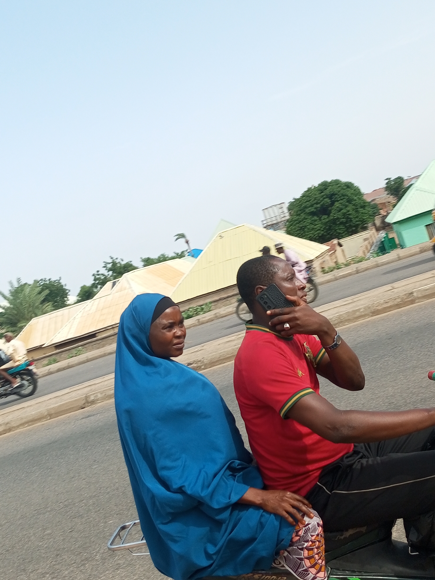 In an outdoor urban environment under a hazy sky, two individuals are riding on a motorcycle. The person in the foreground, a woman wearing a blue hijab and a patterned fabric skirt, is seated behind the driver. The driver, a man wearing a red shirt with green and yellow trim on the collar and black pants, is holding a black smartphone to his right ear, engaged in a conversation. A silver rack is visible on the back of the motorcycle. In the background, a motorcycle with a single rider is moving along a road lined with buildings that have light-colored, sloped roofs. Trees are visible throughout the scene, and the road surface is asphalt.