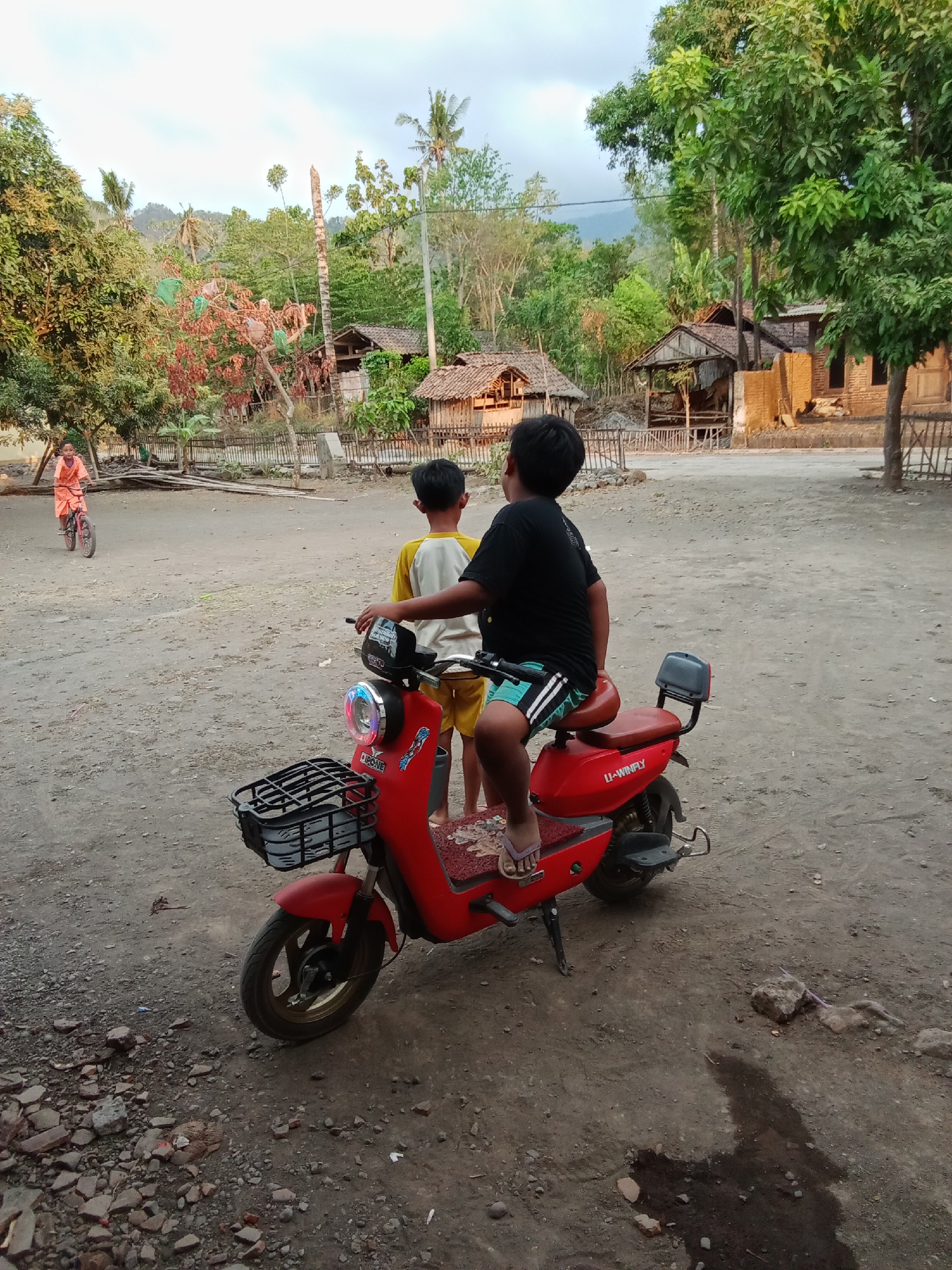 In Selowogo, Indonesia, two young boys are in a dusty, rural area with a red electric scooter. One boy, wearing a black t-shirt and striped shorts, sits on the scooter, facing away from the camera. The other boy, in a yellow and white shirt and yellow shorts, stands next to him. In the background, a younger child rides a bicycle. Several rustic structures with thatched roofs are visible, surrounded by lush green trees and hills. The ground is unpaved and strewn with pebbles and debris. The sky is overcast.