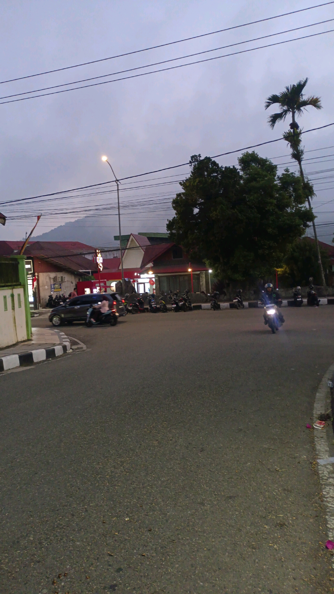 The image depicts a street scene in Padang Panjang, Indonesia, likely during twilight or early evening. In the foreground, a paved road curves to the right, with a white and black striped curb on the left. Parked along the curb are numerous motorcycles and a dark-colored sedan. A motorcycle with its headlights on is in motion in the mid-ground, to the right of the frame, ridden by a person wearing a helmet. Behind the parked vehicles, there are several buildings with red-tiled roofs. A tall palm tree and a large, leafy tree are visible on the right side of the image. Power lines crisscross the sky, and a streetlamp is illuminated on the left. The sky is overcast and a pale grey. The overall impression is of a busy urban street in the late day.