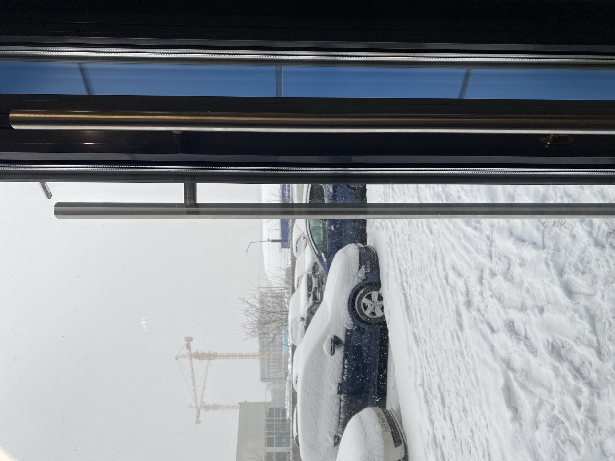 The image, likely taken indoors looking out through a window, depicts a snowy scene. Outside, several cars are parked and covered in snow. A construction crane is visible in the background, along with some buildings. The location is likely Yekaterinburg, Russia, judging by the common architectural and infrastructural elements. The view is framed by the window structure, which includes handles and panes. The lighting suggests daylight.